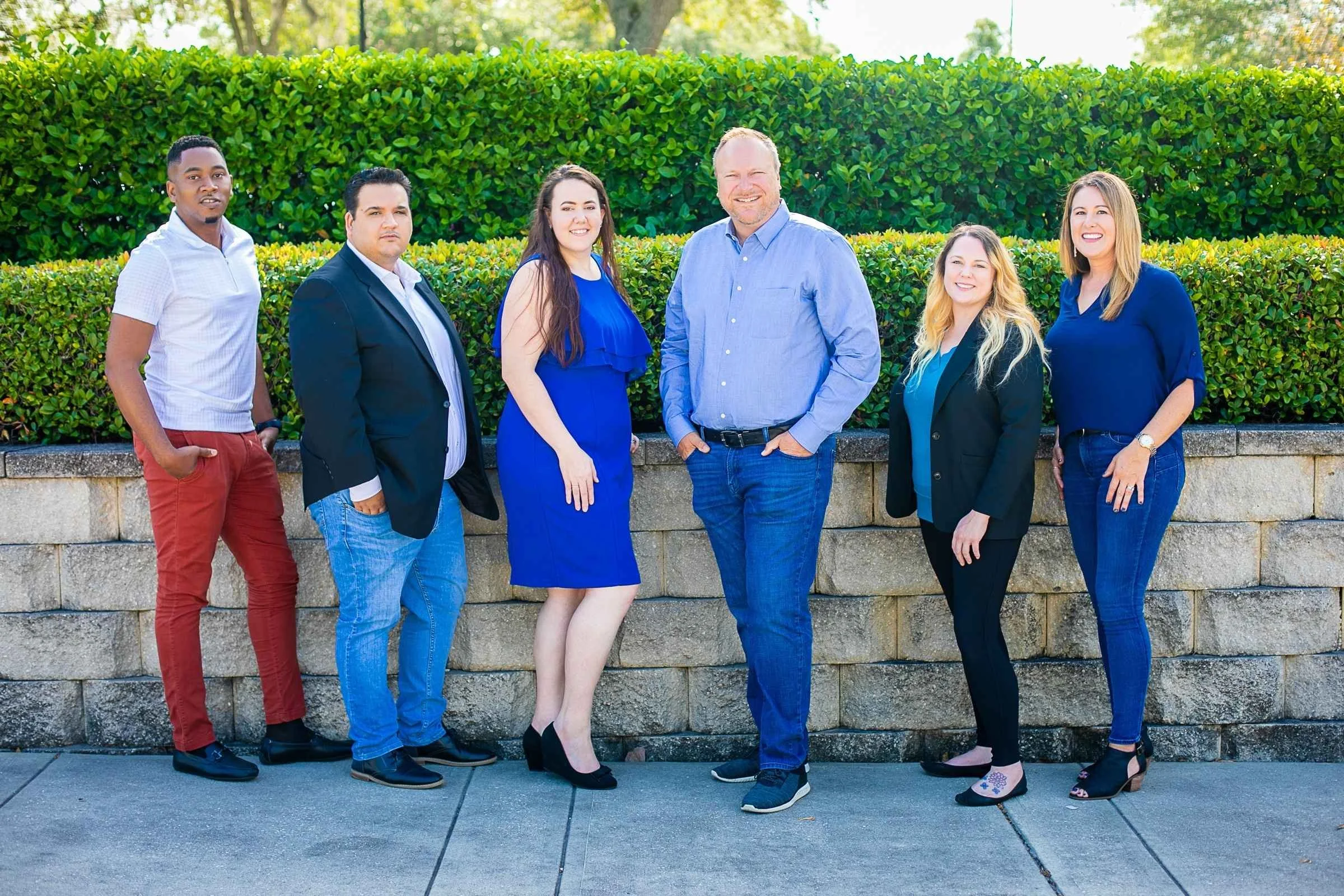 Group of six diverse adults standing outdoors in front of a green hedge, dressed in business casual attire, posing for a team photo by Cannonfire Photography.