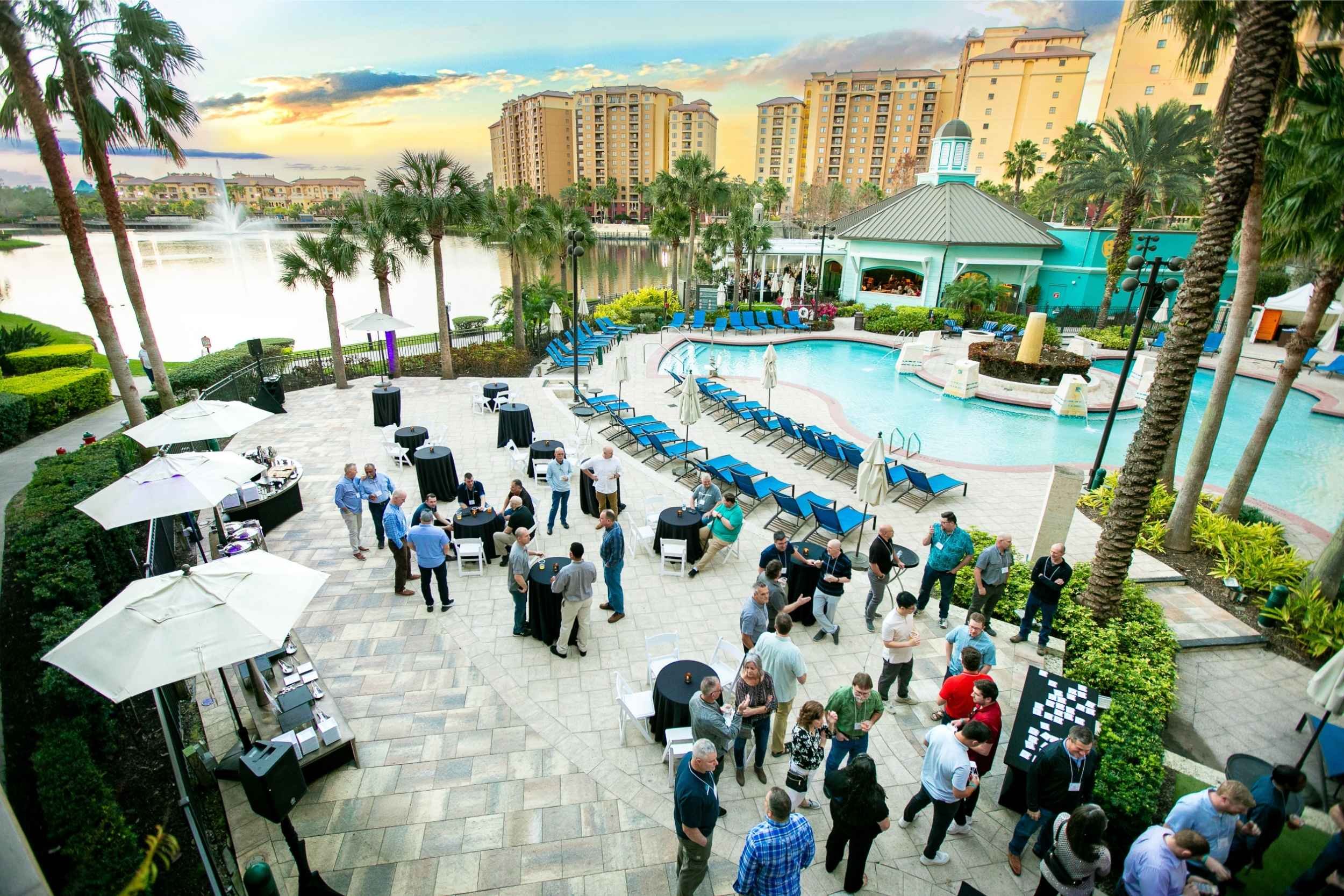An event taking place by a swimming pool with palm trees, high-rise buildings in the background, and a gazebo. People are standing in groups, talking, with tables and umbrellas around.
