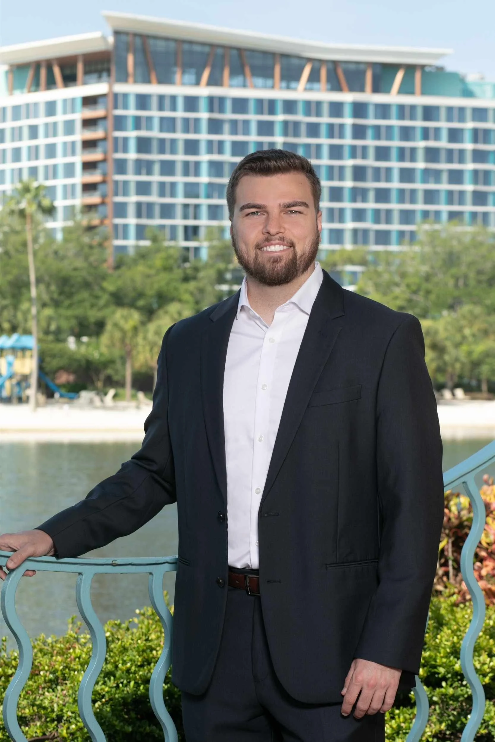 A man in a black suit and white shirt standing outdoors near a railing with a cityscape and river in the background for a headshot with Cannonfire Photography.