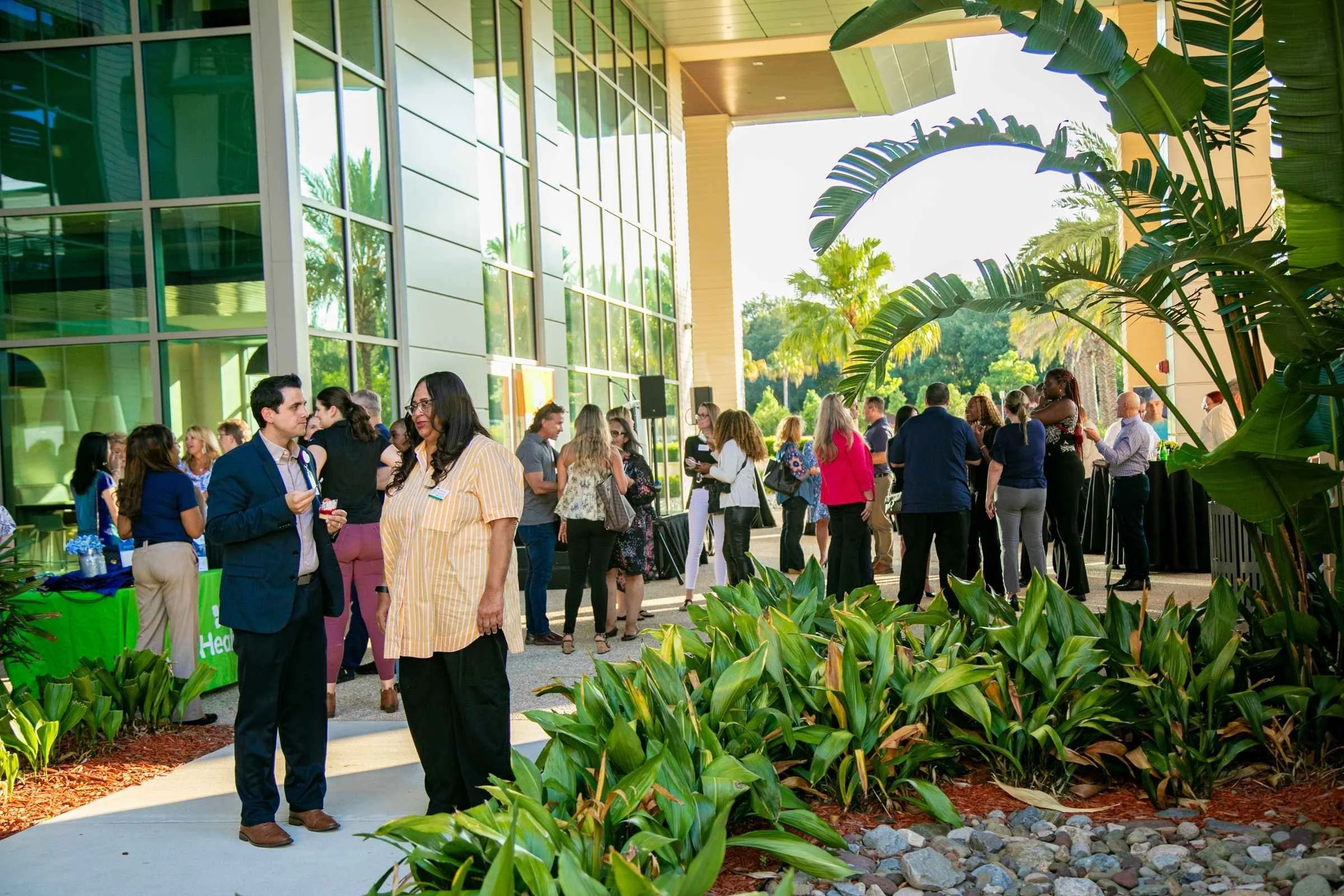People gathered outside a modern building with large glass windows, engaging in conversations during a corporate event, with lush green trees and plants in the surrounding area.