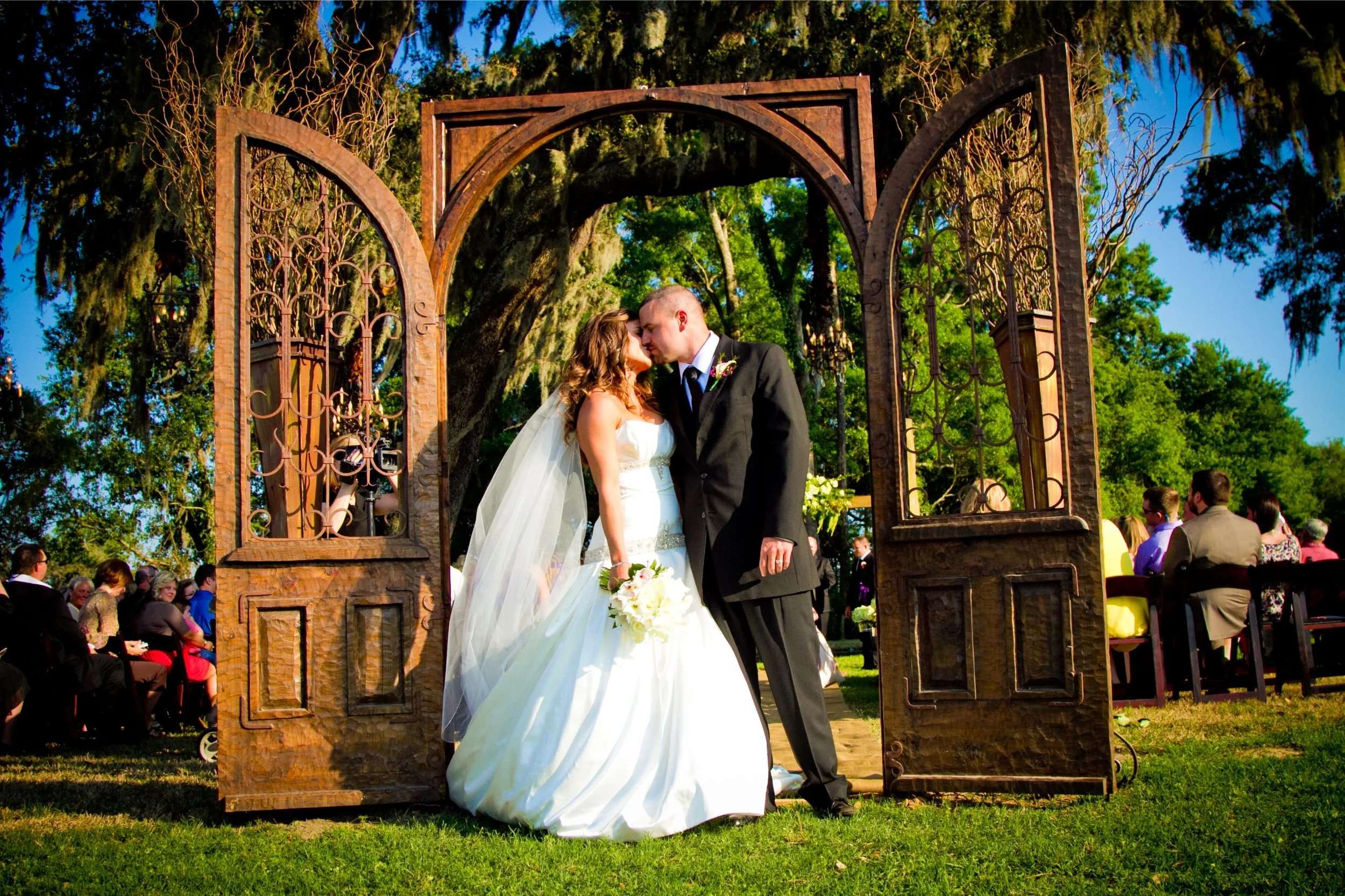 A newlywed couple sharing a kiss under a decorative wooden arch during their outdoor wedding ceremony on a sunny day. Guests are seated in the background surrounded by lush green trees at a wedding photographed by Cannonfire Photography.
