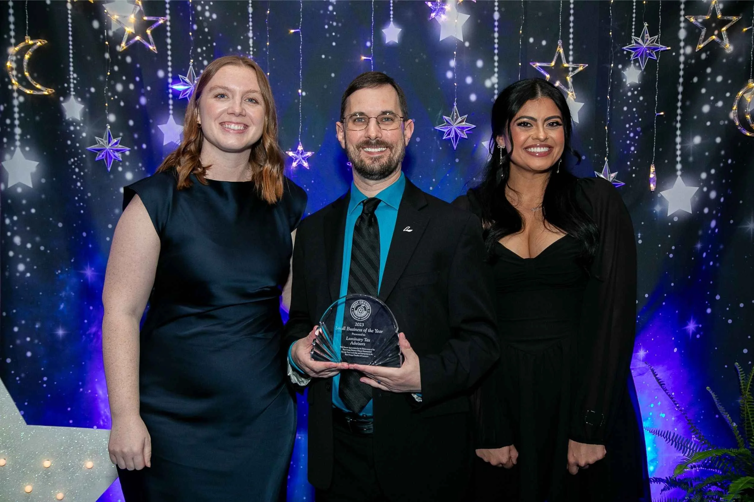 Three people, two women and one man, smiling and standing together at an award ceremony, with festive star and moon decorations and string lights in the background. The man is holding a clear award plaque.