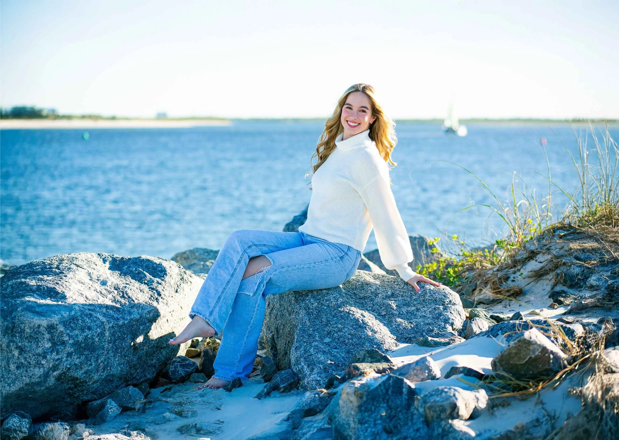 A young woman with blonde hair, wearing a white sweater and ripped jeans, sitting on rocks by a body of water, smiling at the camera on a sunny day for a senior portrait session with Cannonfire Photography.