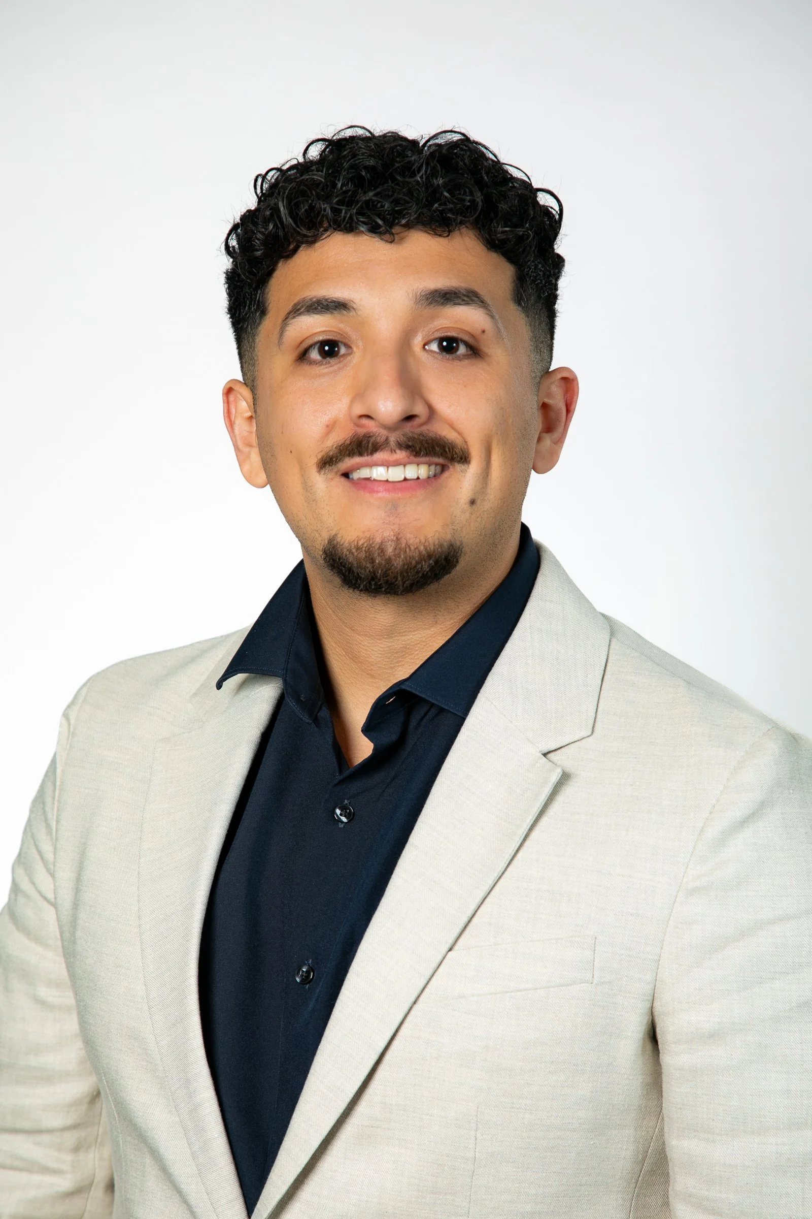 Professional portrait of a young man with curly dark hair, a goatee, smiling, wearing a light-colored blazer and dark shirt against a white background for a headshot with Cannonfire Photography in Orlando.