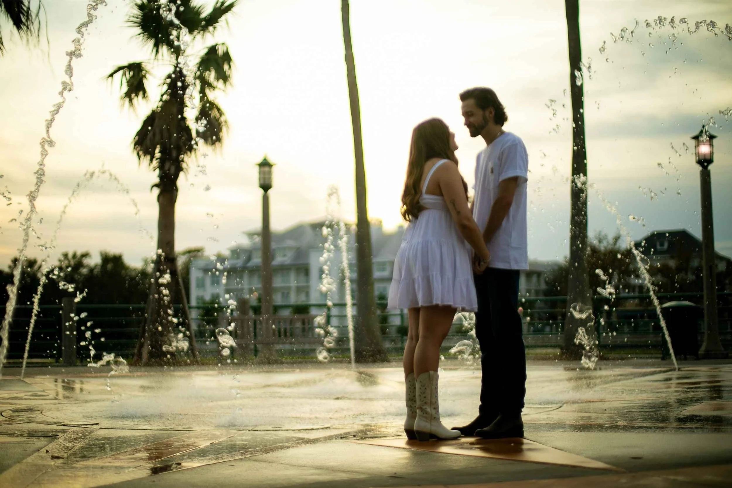 A couple stands close together, holding hands, in front of a fountain at sunset. The woman wears a white dress and cowboy boots, and the man wears a white t-shirt and dark jeans. Palm trees and a building are in the background.