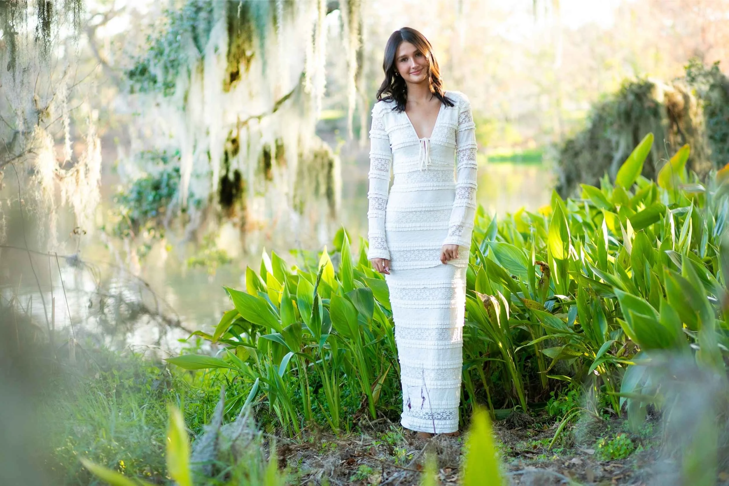 Woman in a white dress standing among green plants by a waterway in a lush natural setting for a senior portrait session with Cannonfire Photography.