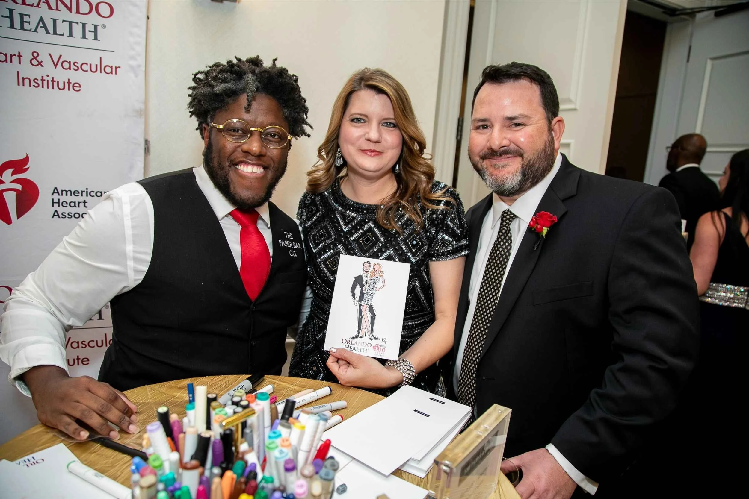 Three people posing for a photo at a health event, with a woman holding a booklet and a table filled with colorful markers in front of them, and a banner reading 'American Heart Association.'