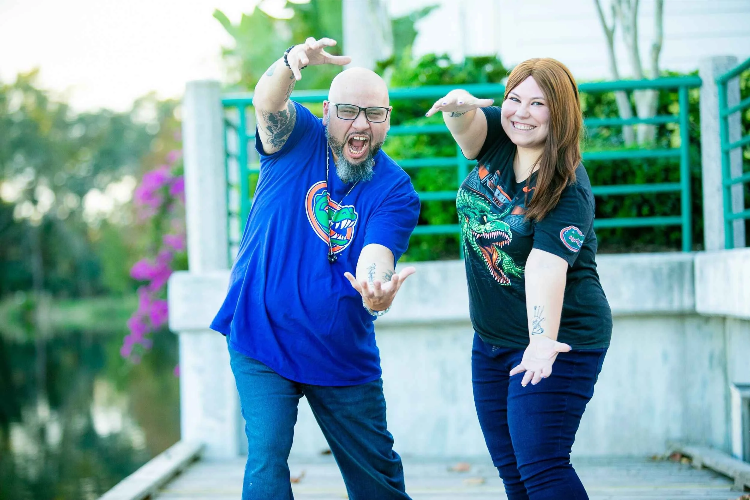 Two smiling people, a man with glasses and a woman with red hair, posing playfully on a bridge outdoors, with greenery and purple flowers in the background for an engagement photography session with Cannonfire Photography.