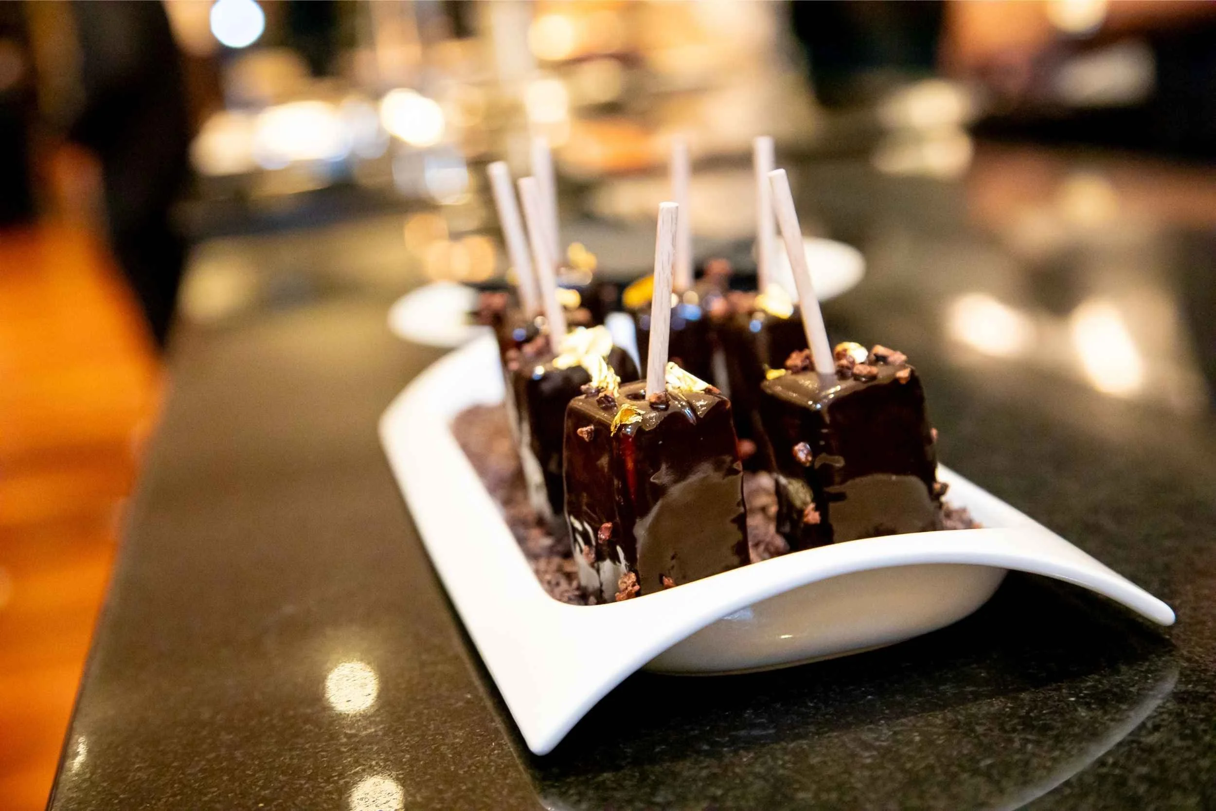Chocolate dessert cubes on sticks with chocolate sauce and toppings, arranged on a white serving dish on a dark countertop