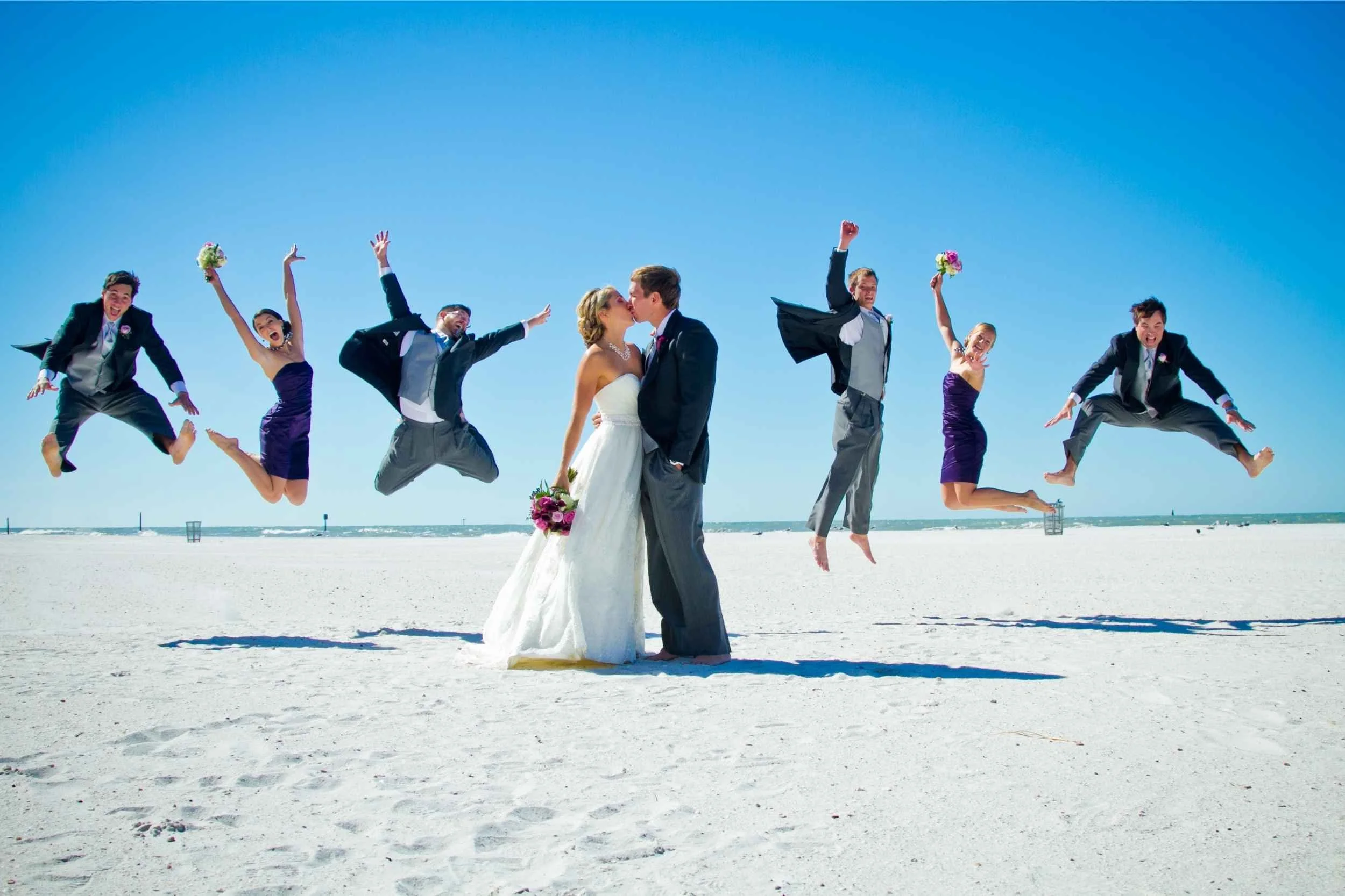 A wedding party on a beach with a clear blue sky, featuring the bride and groom kissing surrounded by friends and bridesmaids jumping and holding bouquets at a wedding photographed by Cannonfire Photography.