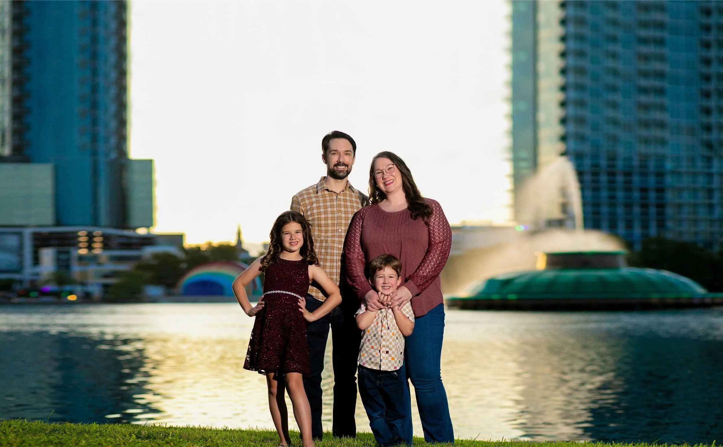 A family of four standing outdoors near Lake Eola with downtown Orlando city buildings in the background at sunset.