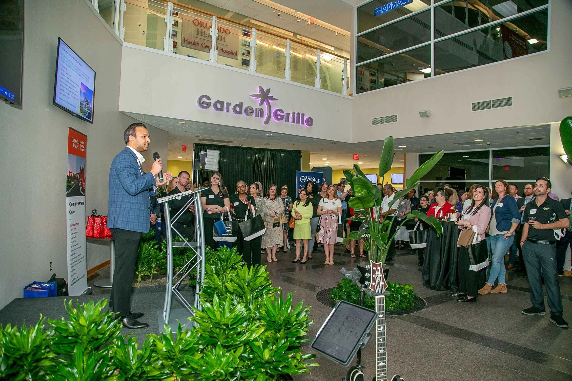 A man in a blue checkered jacket speaks at a podium in a busy indoor event space with a crowd of diverse attendees listening. There are plants in the foreground, and signs for a garden grill restaurant, pharmacy, and TV station in the background.