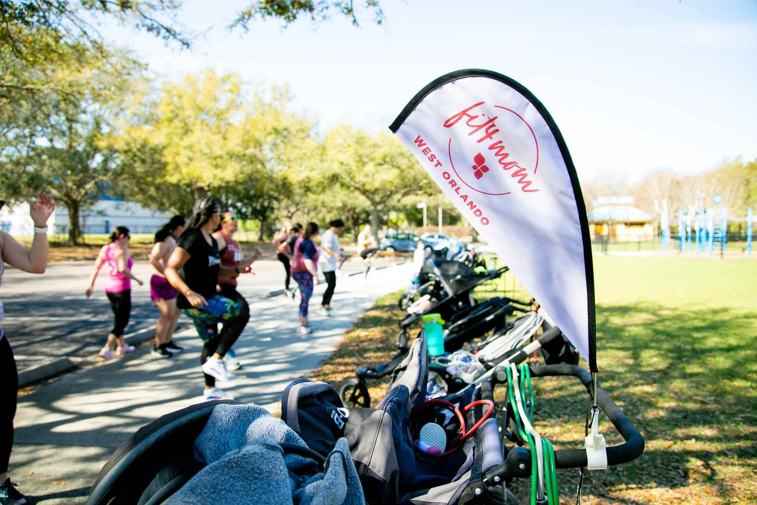 A group of women participating in a fitness activity outdoors in a park, with a row of strollers and fitness equipment in the foreground, and trees and a playground in the background for an advertising photography session by Cannonfire Photography.