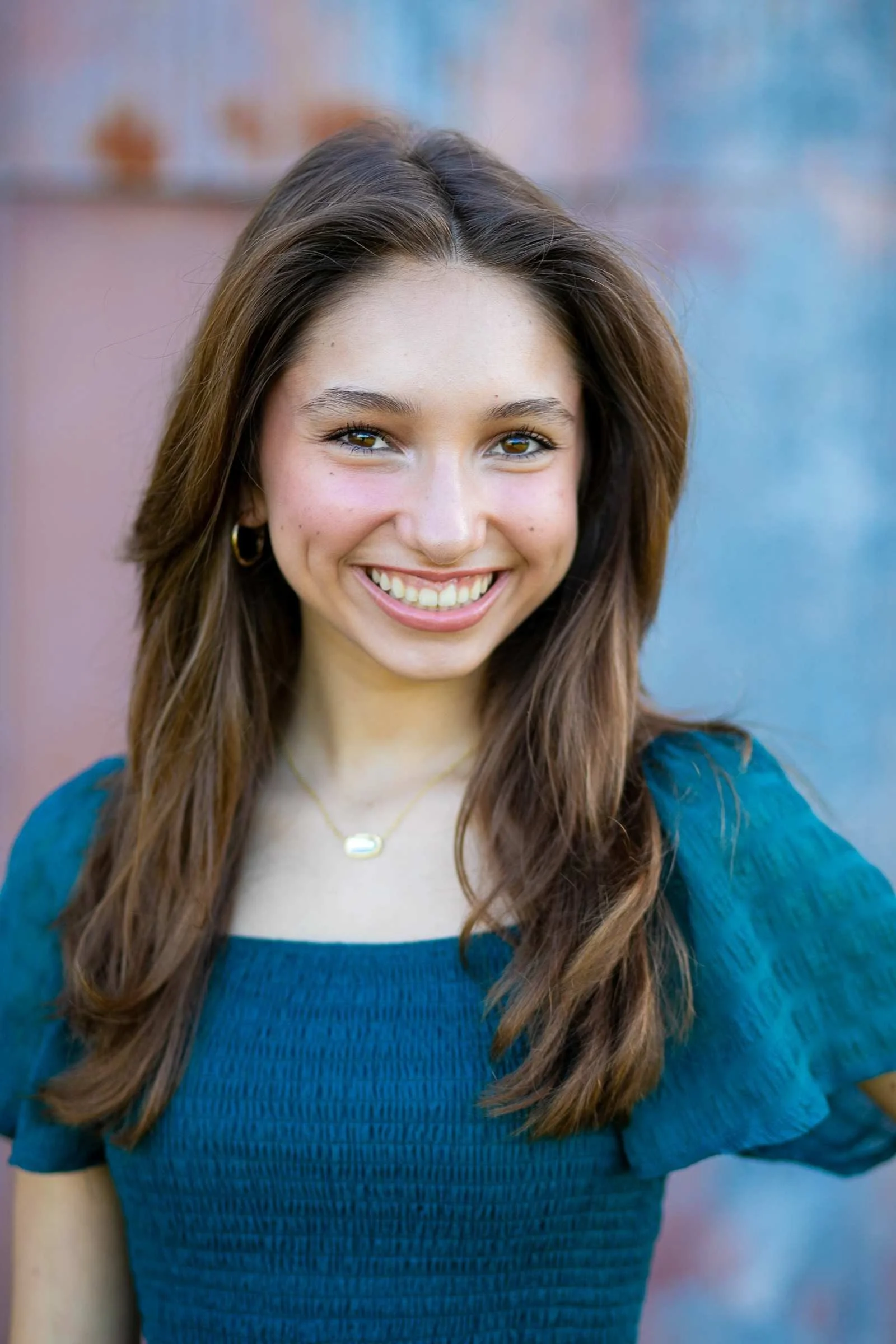 Close-up portrait of a young woman smiling with long brown hair, wearing a blue top and gold jewelry, outdoors with a blurred colorful background during a family portrait session with Cannonfire Photography.