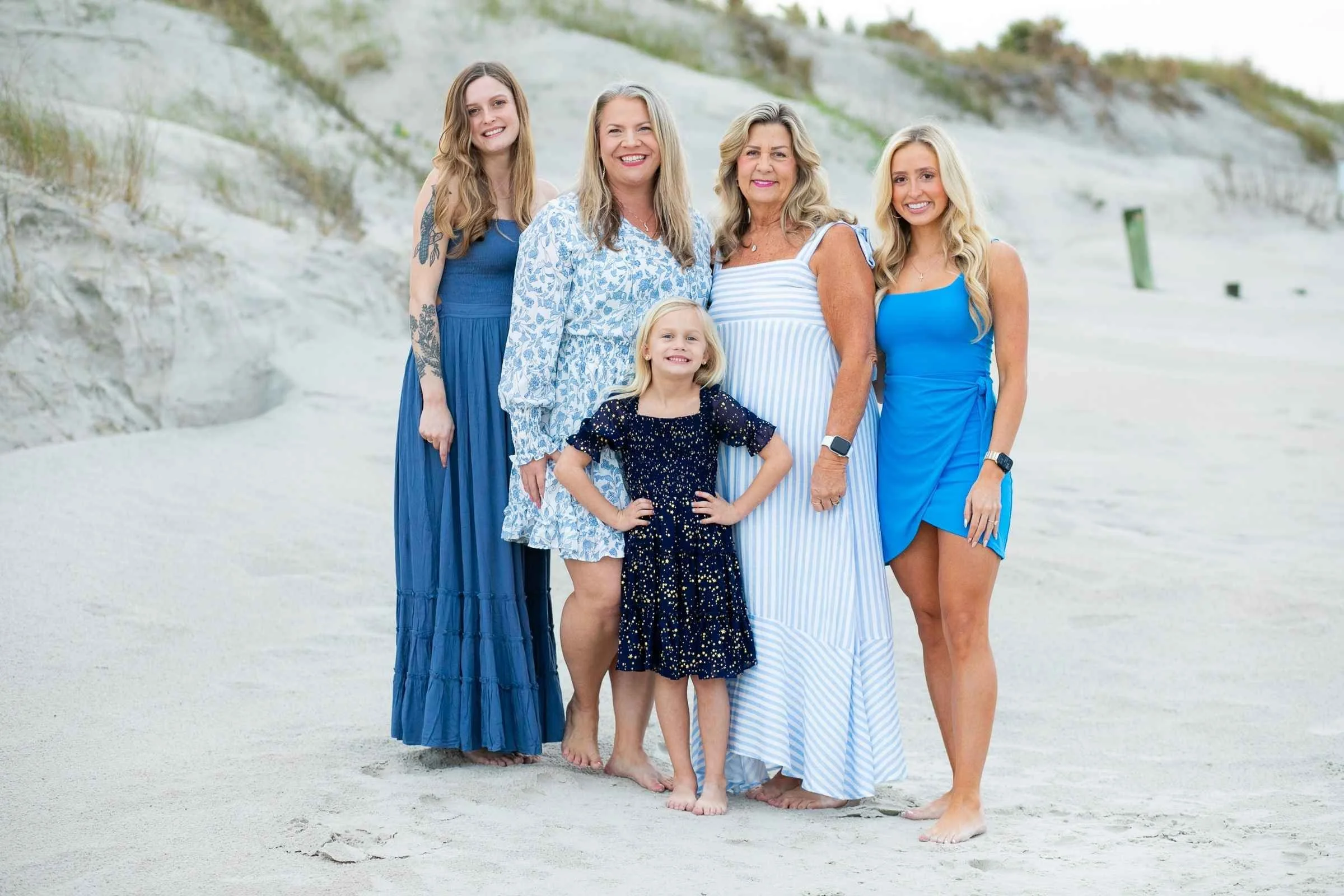 Group of five women and girls standing on a sandy beach, smiling at the camera, with cliffs and greenery in the background. They are dressed in blue and white outfits.