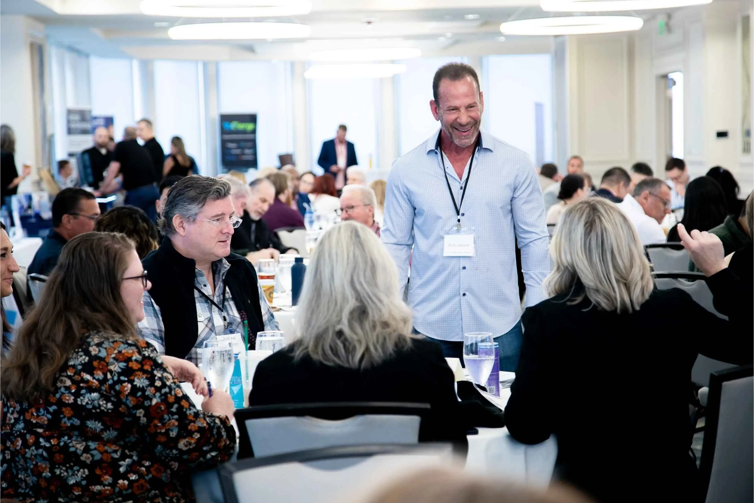 A group of people attending a corporate event in a bright, modern room in Orlando. A man with a lanyard is standing and smiling while talking to seated attendees.
