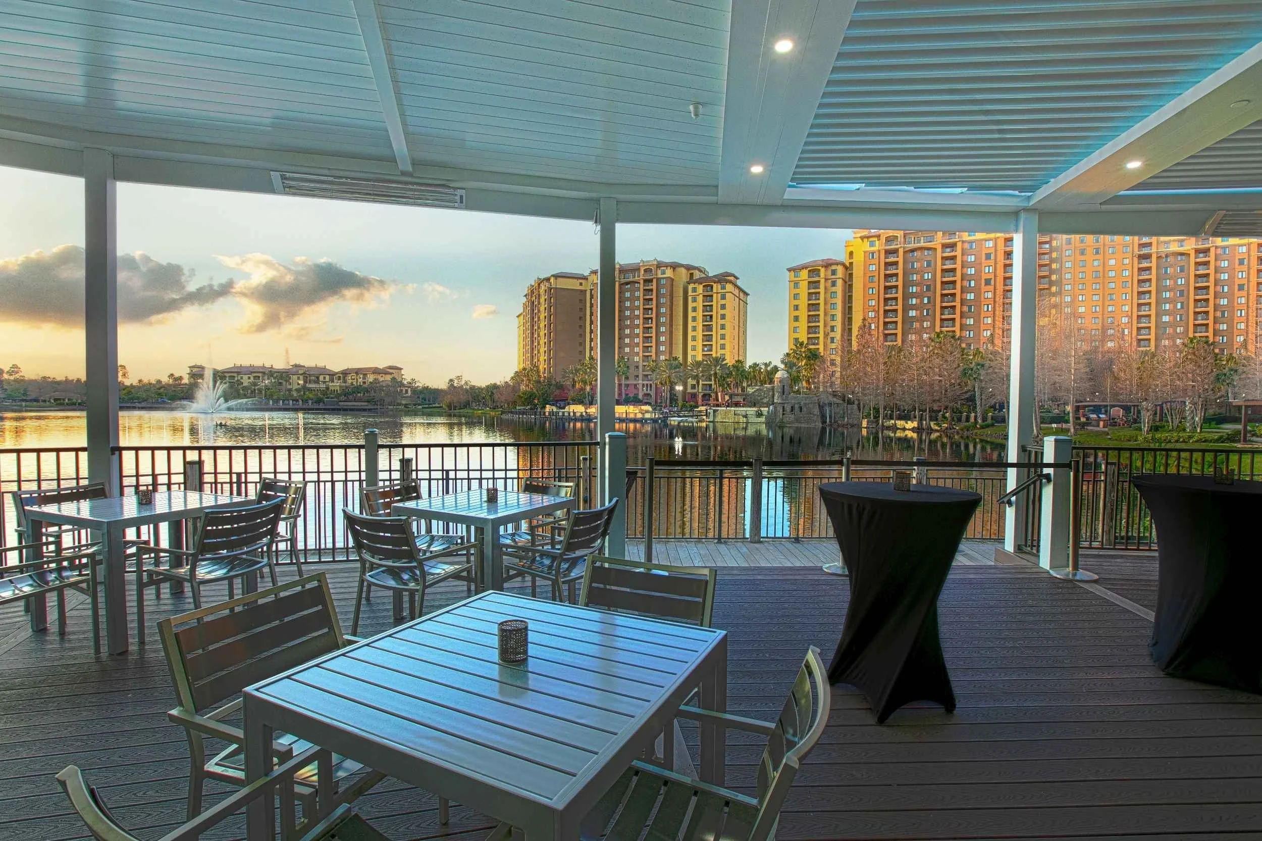 Outdoor lakeside dining area with tables and chairs, overlooking a lake with a fountain and multi-story hotel buildings in the background during sunset.