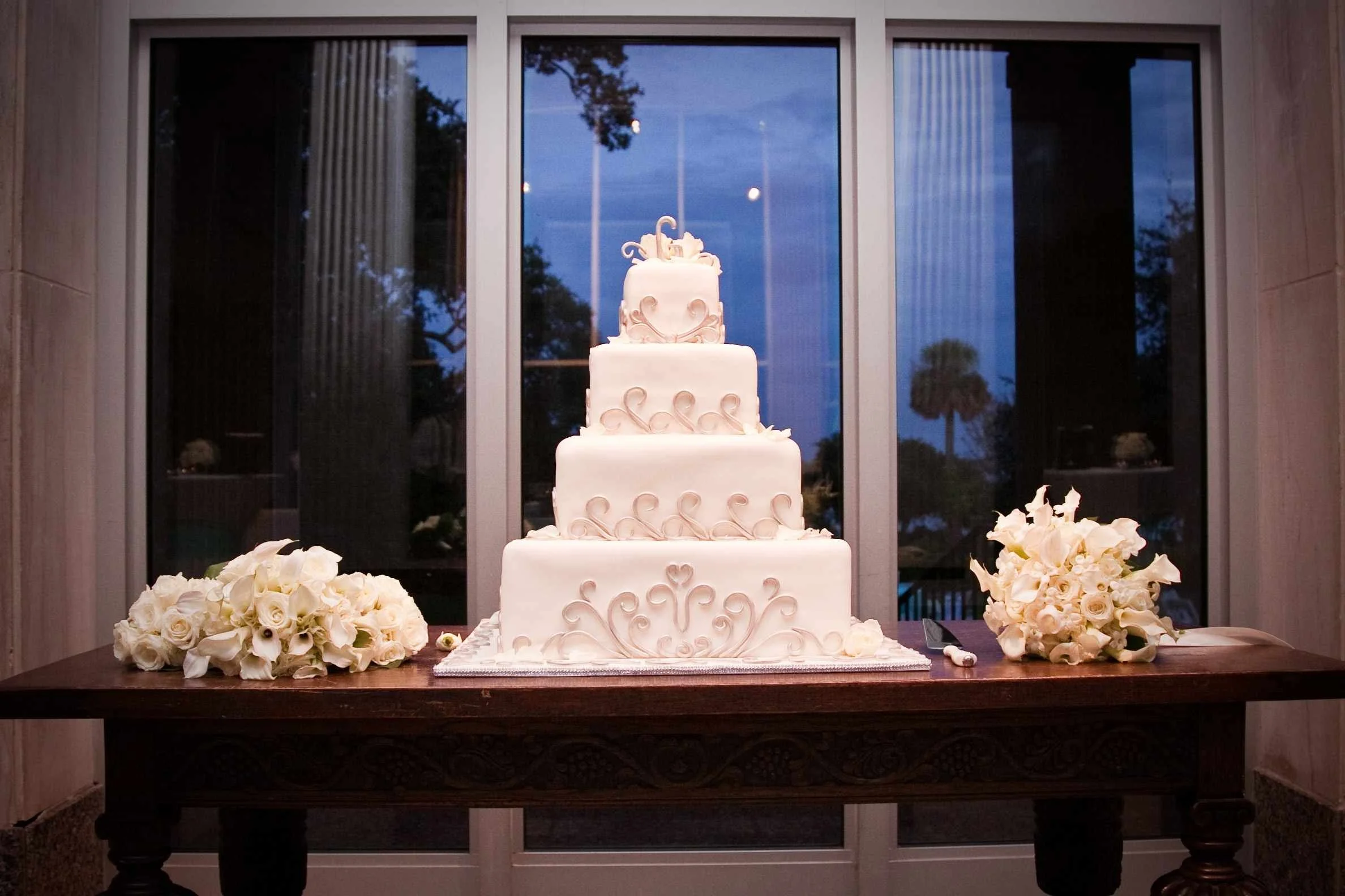 A multi-tiered white wedding cake decorated with swirl details, placed on a table with bouquets of white roses and greenery surrounding it, in front of a large window at a wedding photographed by Cannonfire Photography.