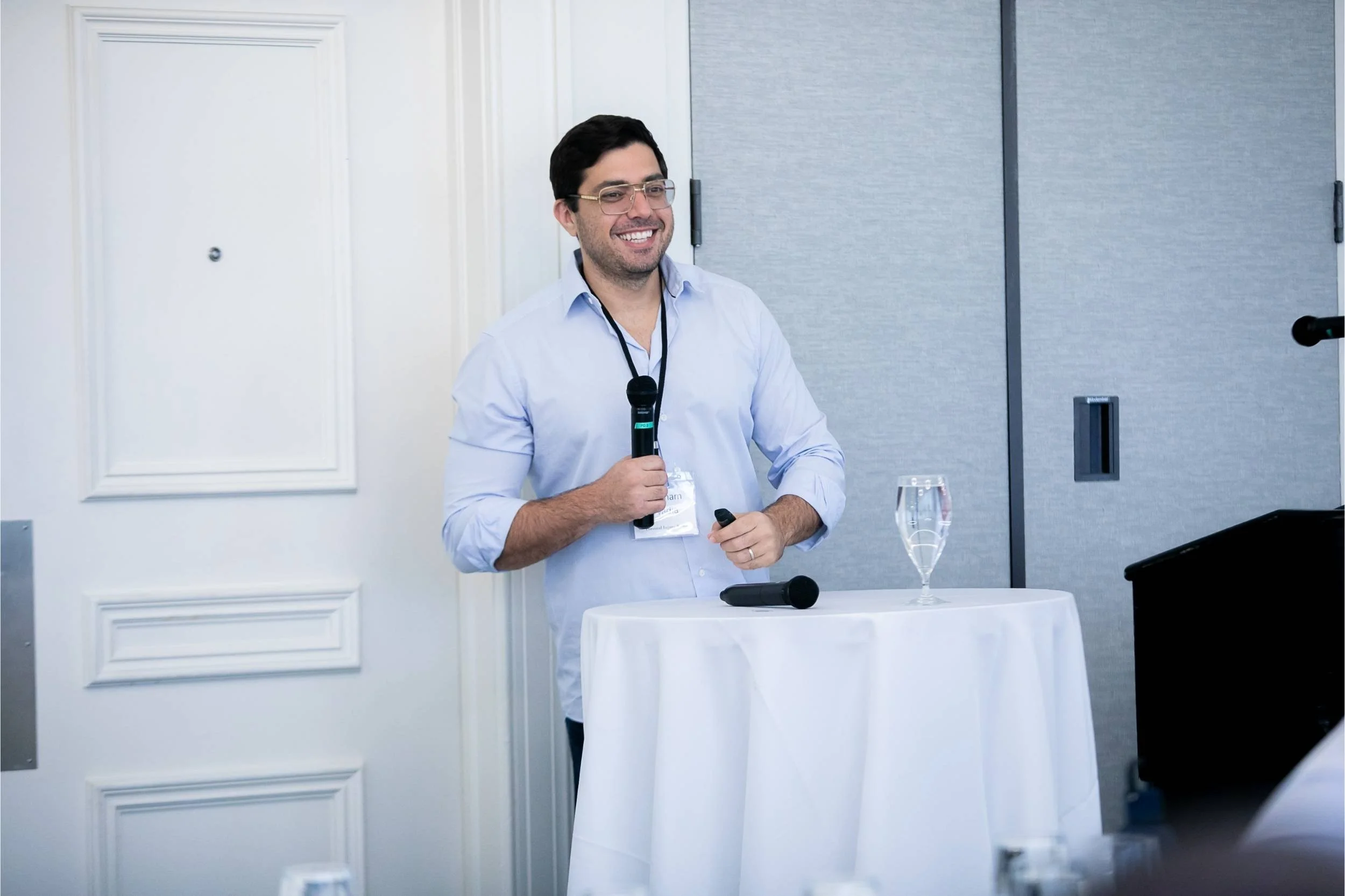 A man with dark hair and glasses, wearing a light blue shirt, is smiling and holding a microphone and clicker at a presentation or conference, standing behind a round table with a glass of water.