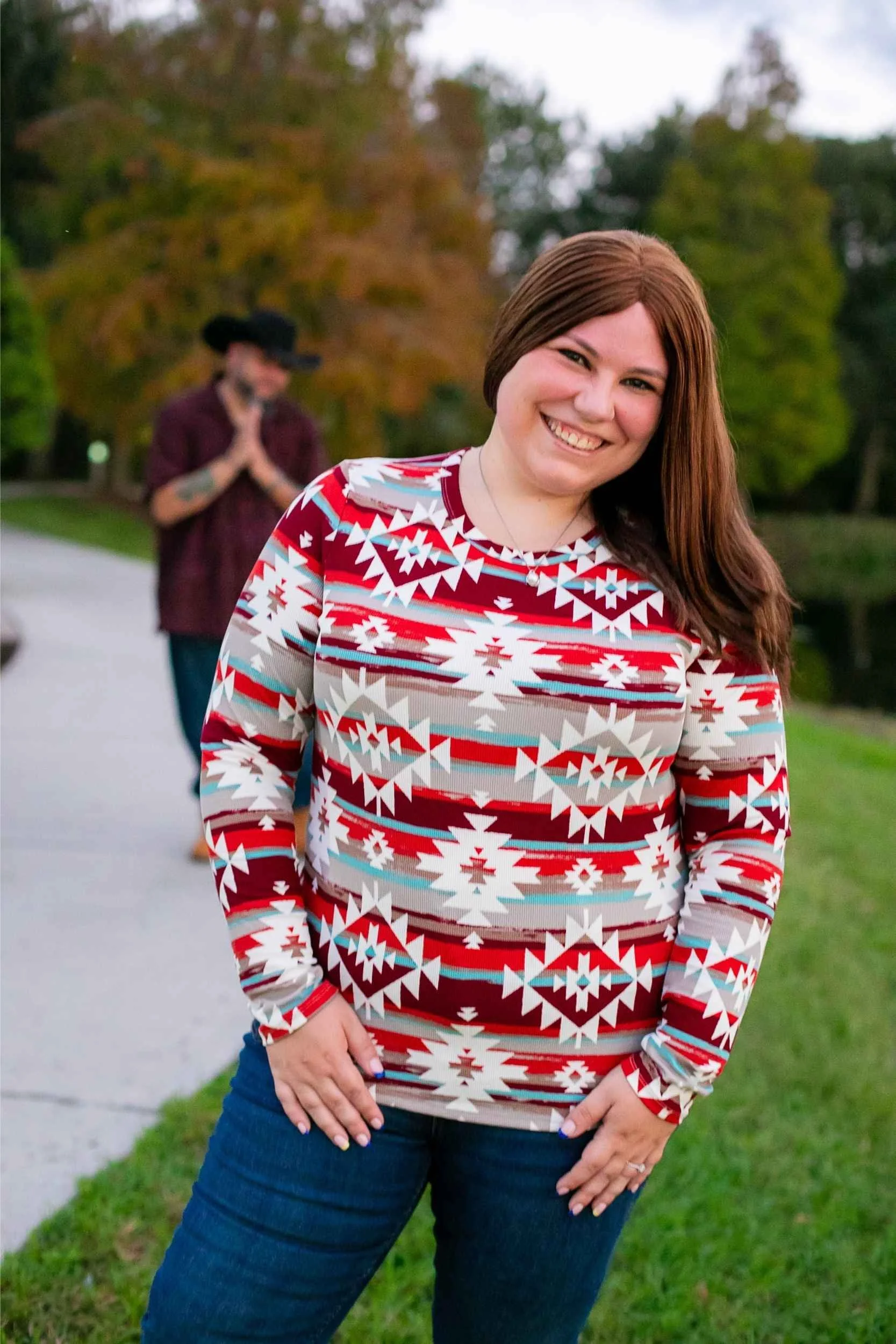 A smiling woman with long brown hair wearing a colorful, patterned sweater standing outdoors near a body of water with lush green and orange trees in the background. A person with a hat and tattoos is in the blurred background.
