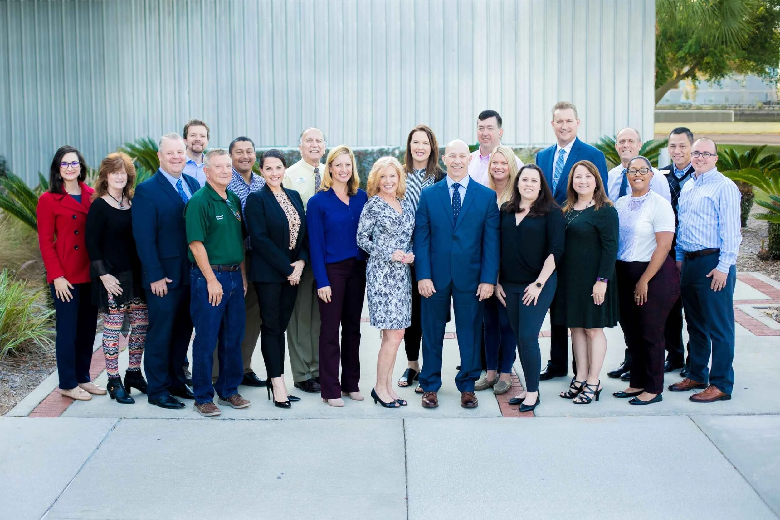 Group of professionally dressed men and women standing outdoors in front of a corrugated metal building and green plants, smiling at the camera for a team photo by Cannonfire Photography.