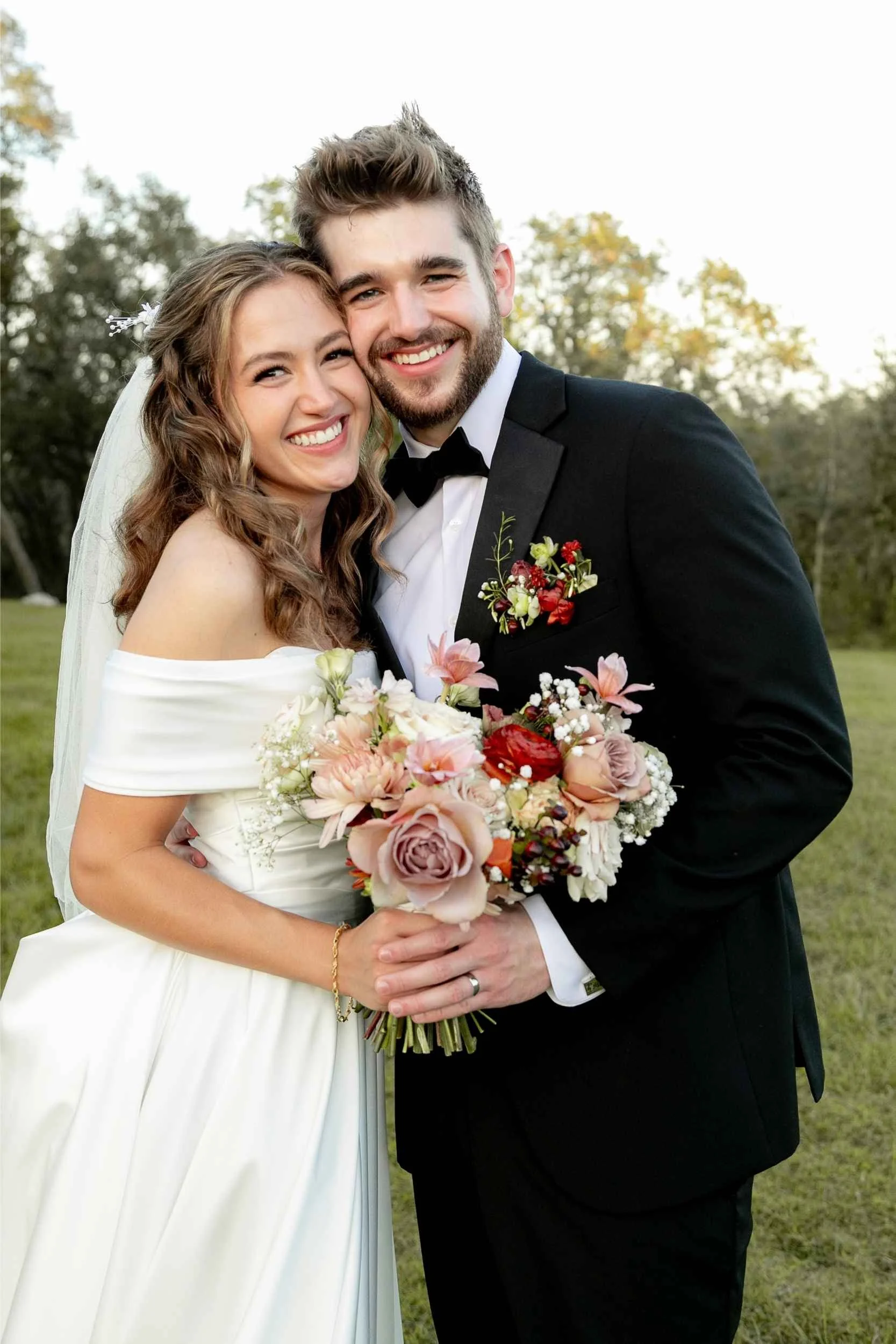 A newlywed couple smiling and embracing outdoors. The bride is wearing a white off-shoulder wedding dress and holding a bouquet of pink, red, and white flowers. The groom is dressed in a black tuxedo with a white shirt and black bow tie, holding the 