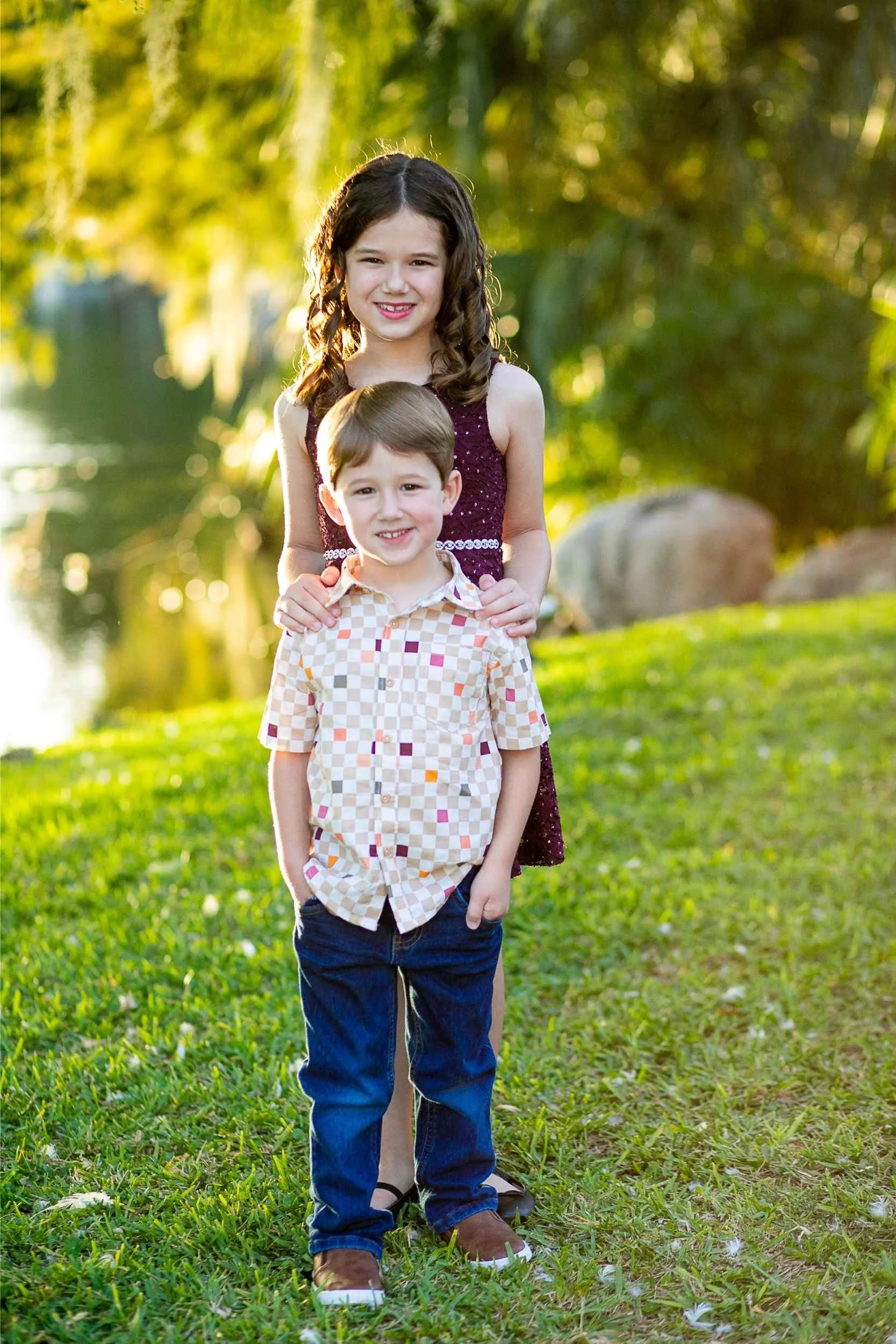 A girl and a boy standing outdoors on a grassy area near a body of water, with trees and rocks in the background, during golden hour sunlight during a family portrait session with Cannonfire Photography.
