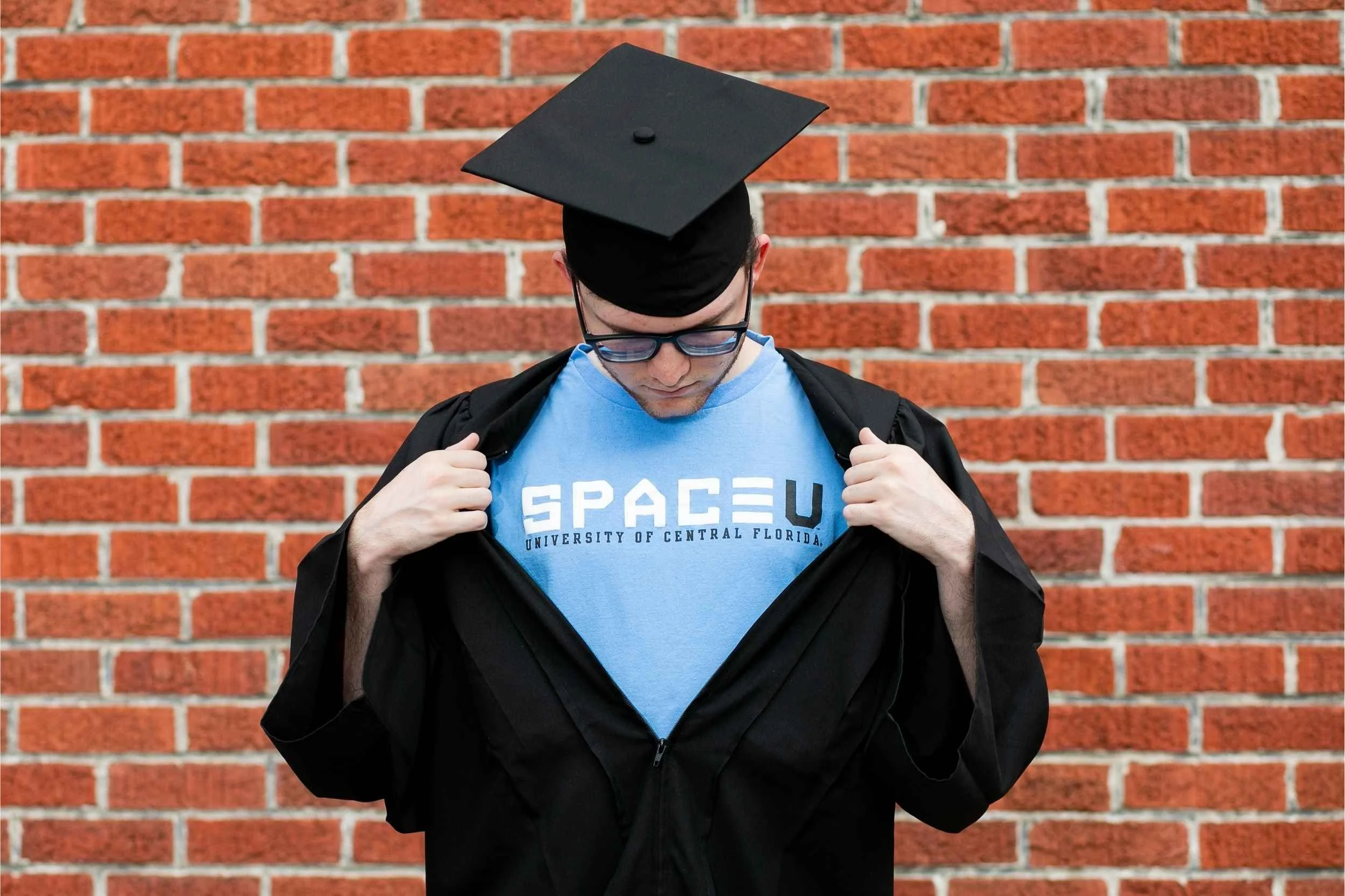 Young man in graduation cap and black gown opening his blue T-shirt that says 'SPACEU' with smaller text 'UNIVERSITY OF CENTRAL FLORIDA,' standing against a brick wall for a senior portrait session with Cannonfire Photography.