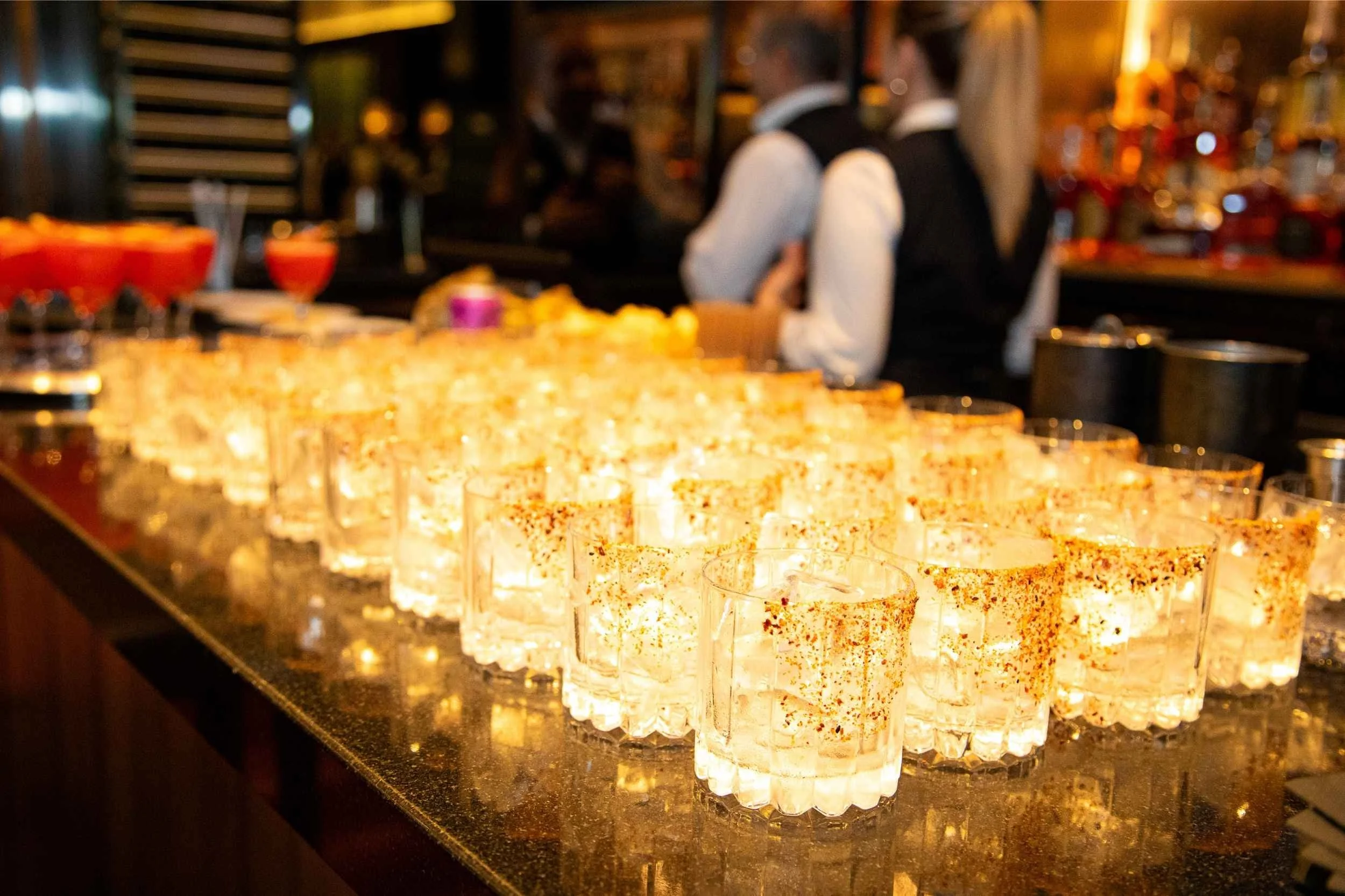 A row of shot glasses filled with clear liquid and sprinkled with red spices on a dark bar counter, with two bartenders in black vests and white shirts working in the background.