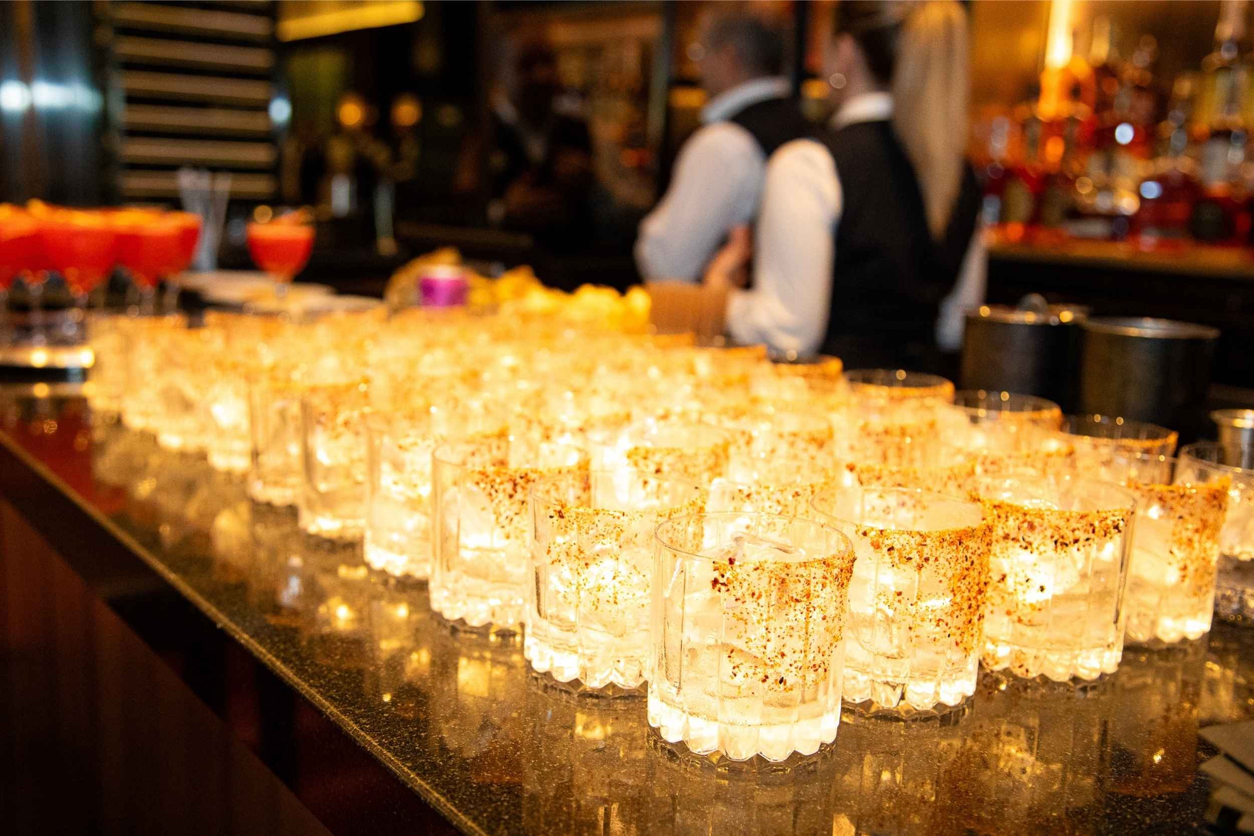 A row of shot glasses filled with clear liquid and sprinkled with red spices on a dark bar counter, with two bartenders in black vests and white shirts working in the background.