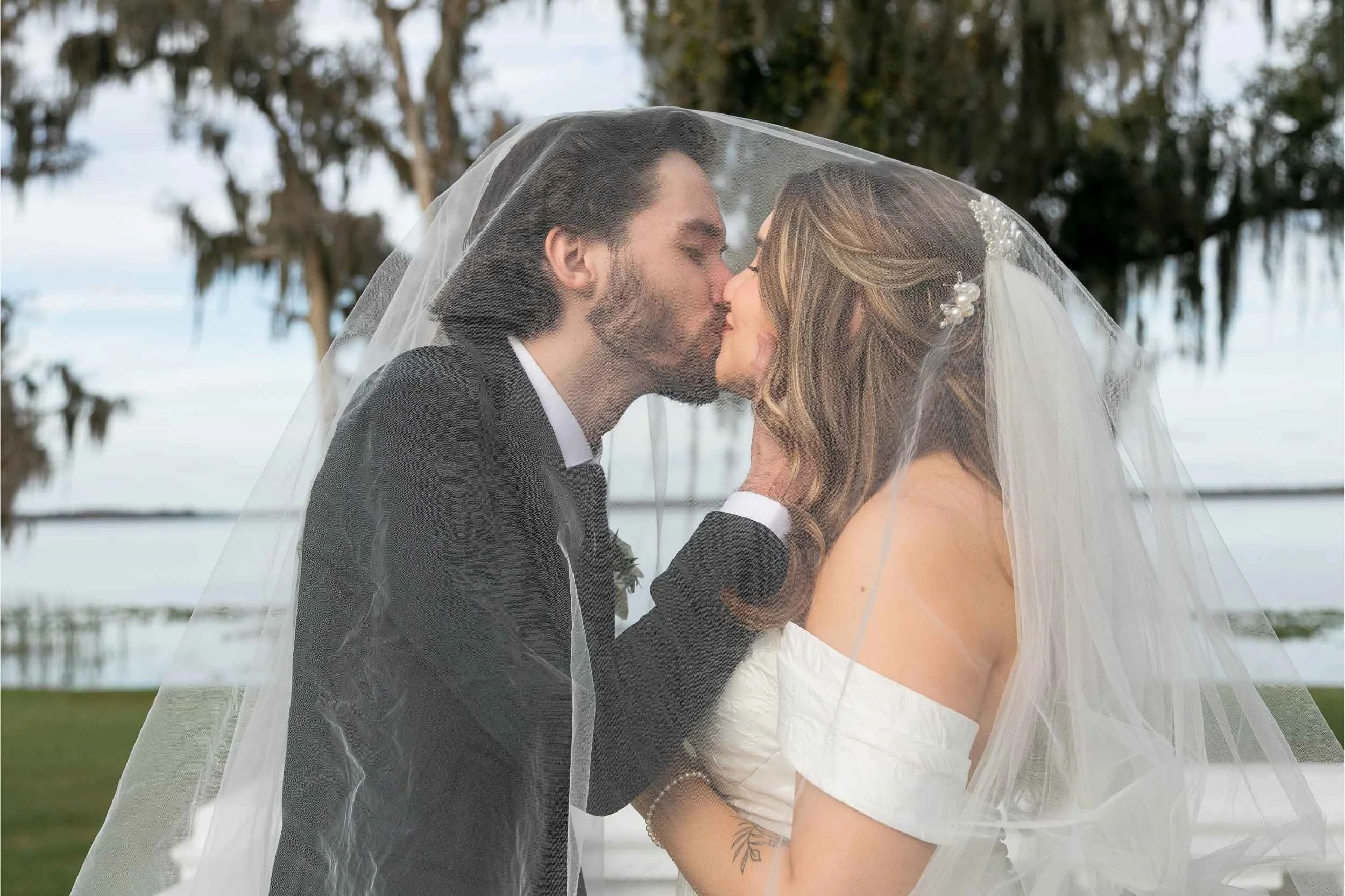 A bride and groom kissing under a wedding veil outdoors near water and trees for a wedding photographed by Cannonfire Photography in Lake Wales.