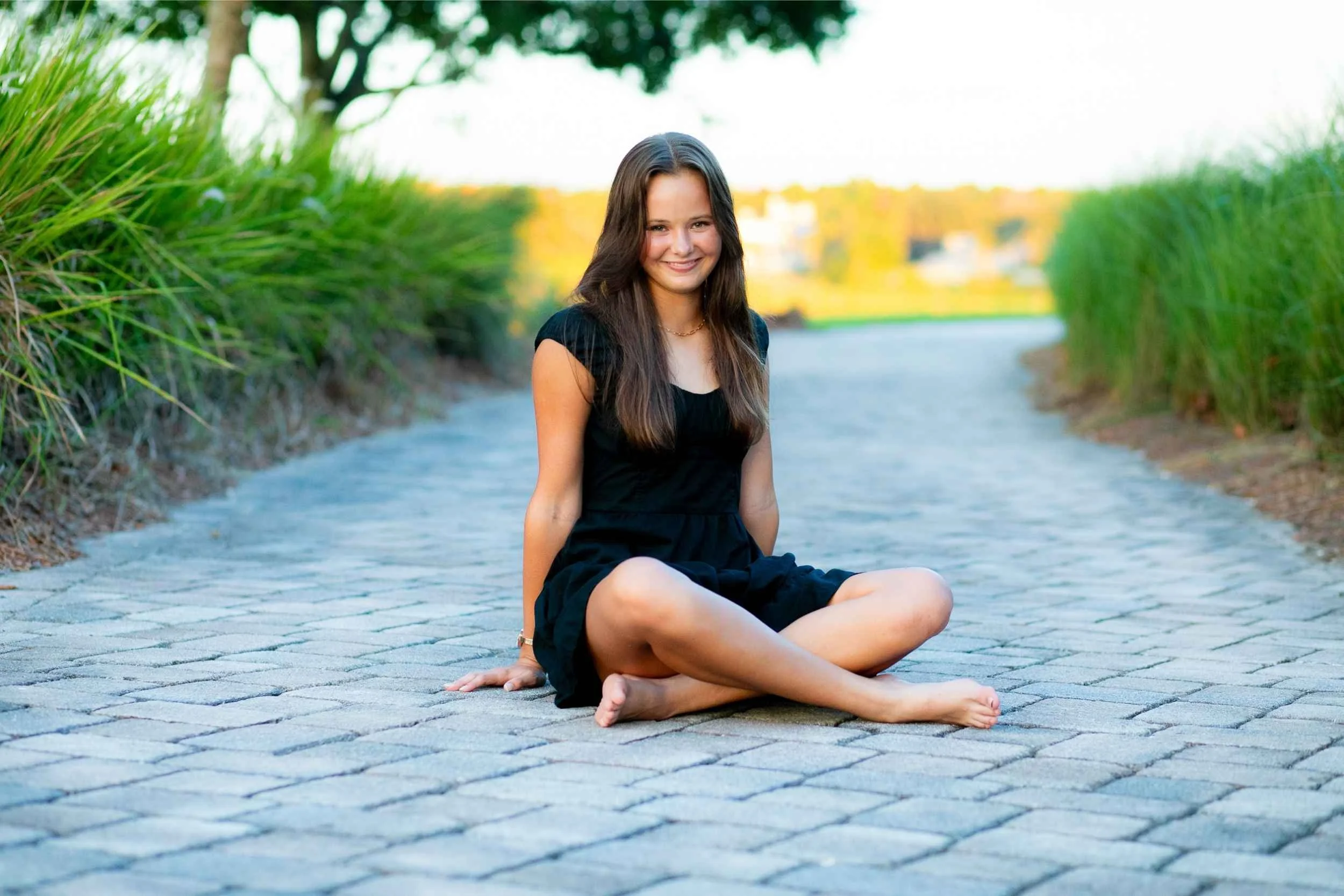 A young woman with long brown hair, wearing a black dress, sitting cross-legged on a paved pathway surrounded by green tall grass and trees, smiling at the camera during daytime for a senior portrait session with Cannonfire Photography.