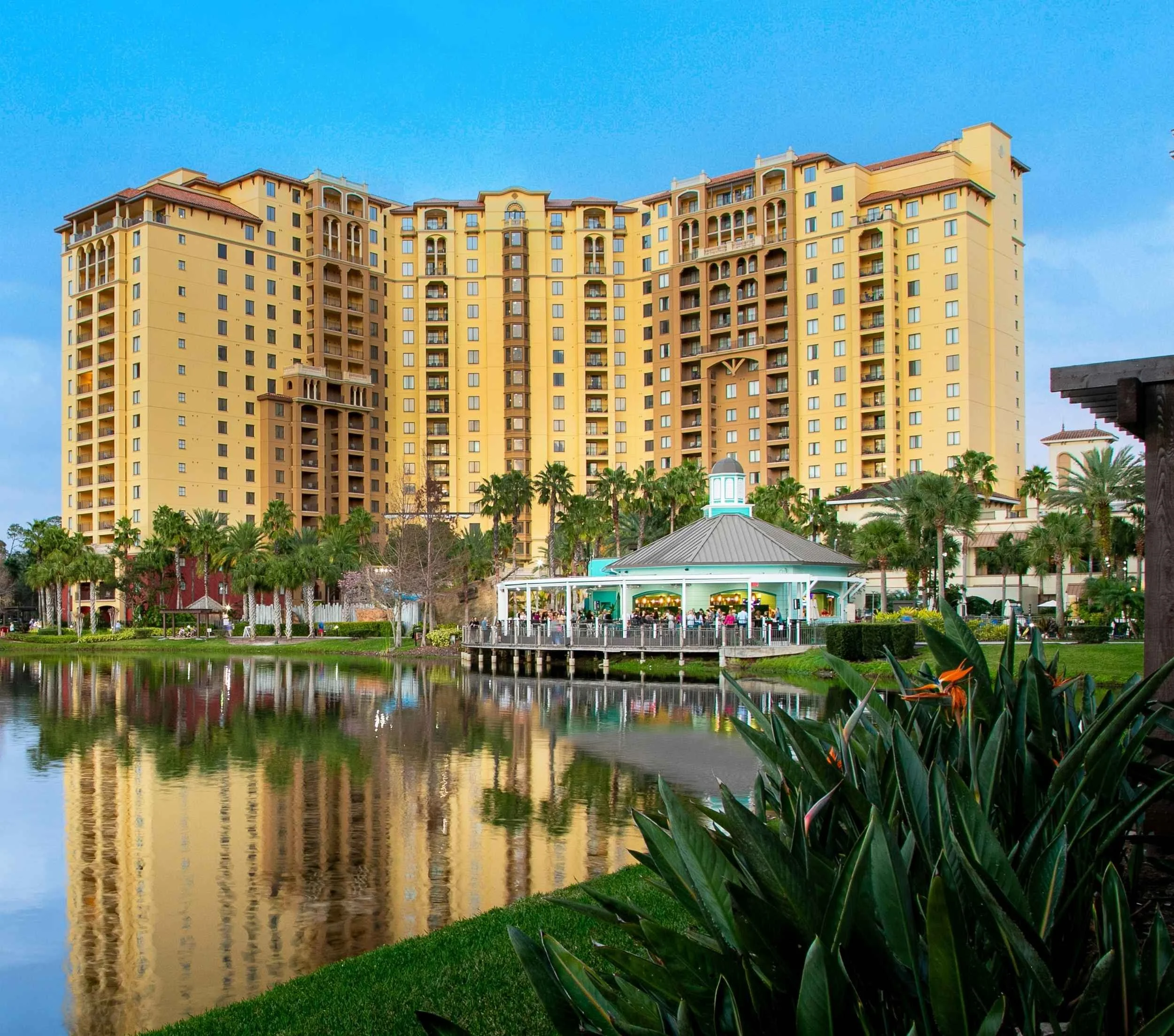 A large hotel with multiple balconies is reflected in a pond in front of it, with a white pavilion and lush palm trees surrounding the area.