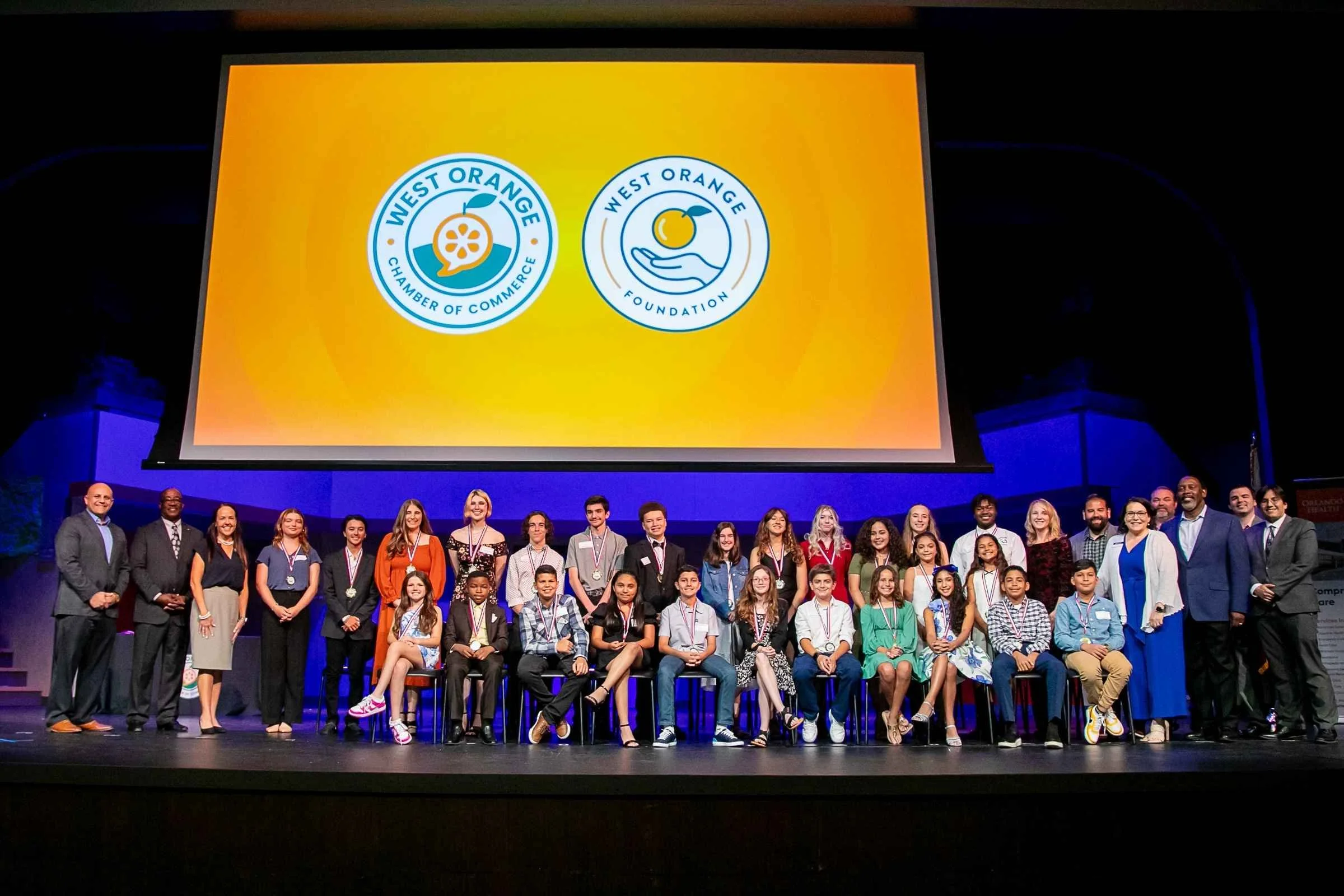Group of diverse children and adults on stage at an event with a large screen displaying the logos of West Orange Chamber of Commerce and West Orange Foundation above them.