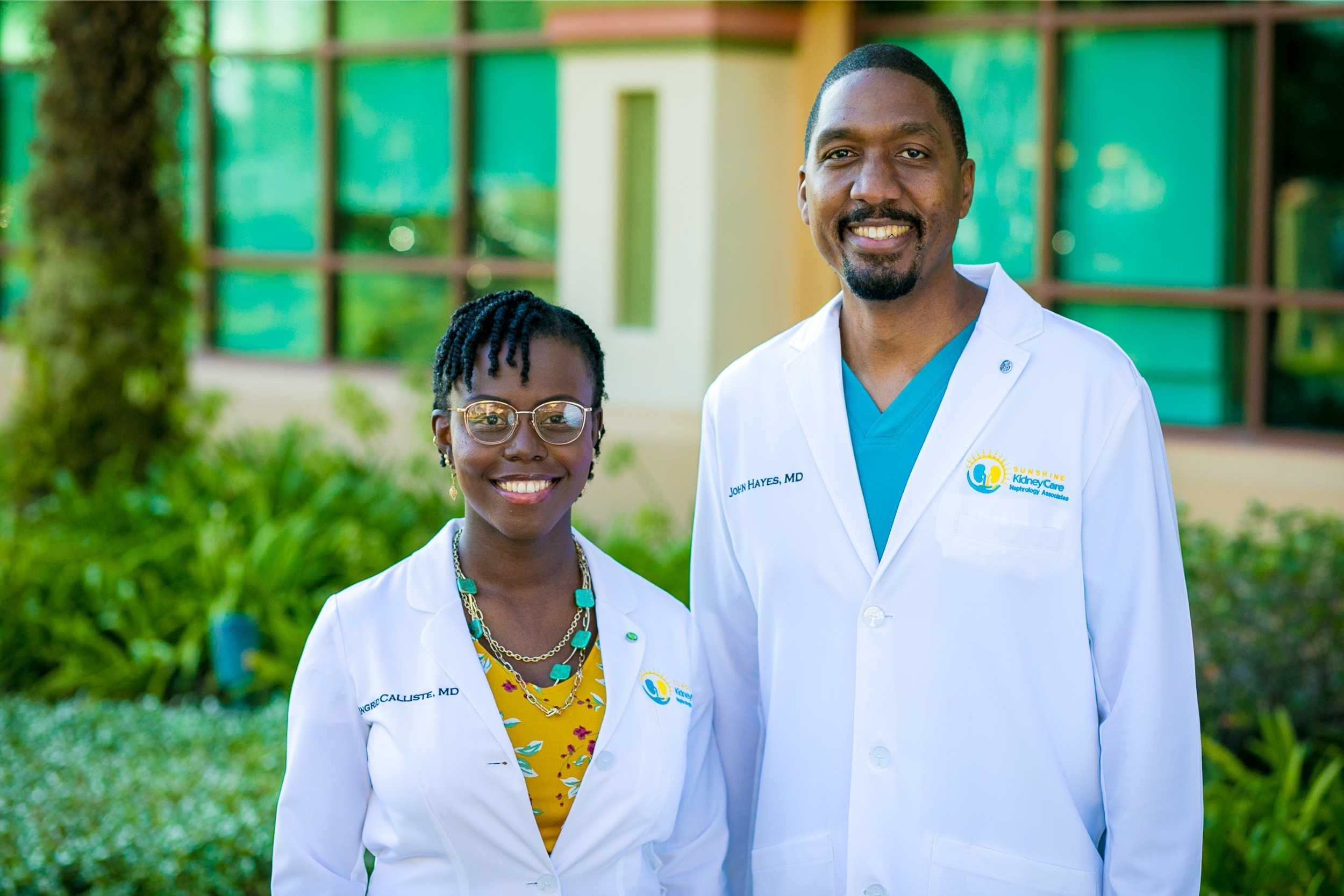 Two medical professionals, a woman and a man, stand outdoors in front of a building with large windows. They are wearing white lab coats with name tags and smile at the camera. The woman has short braided hair, glasses, and colorful accessories. The 