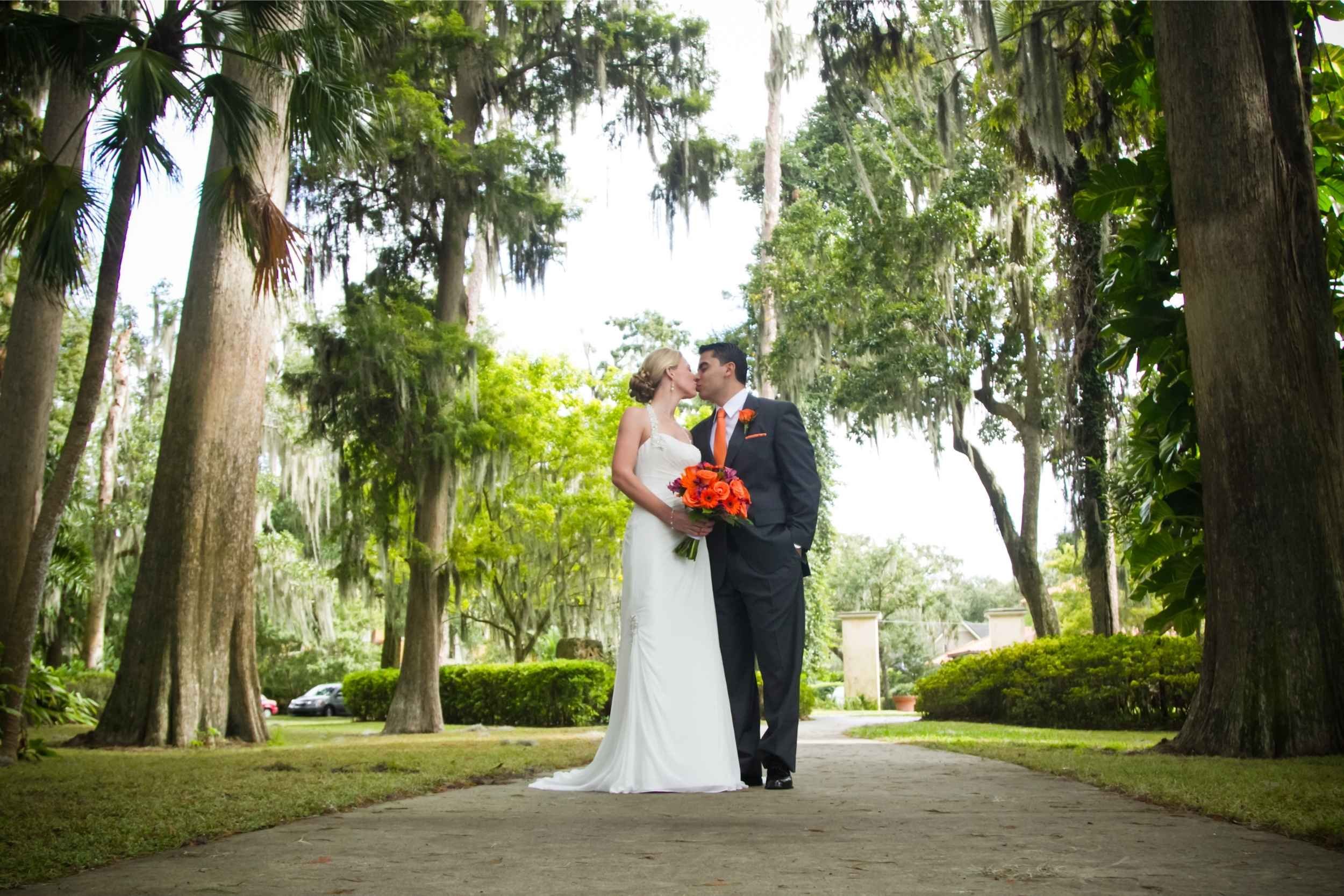 A bride and groom sharing a kiss in a lush, green outdoor setting with tall trees and Spanish moss hanging from the branches at a wedding photographed by Cannonfire Photography.