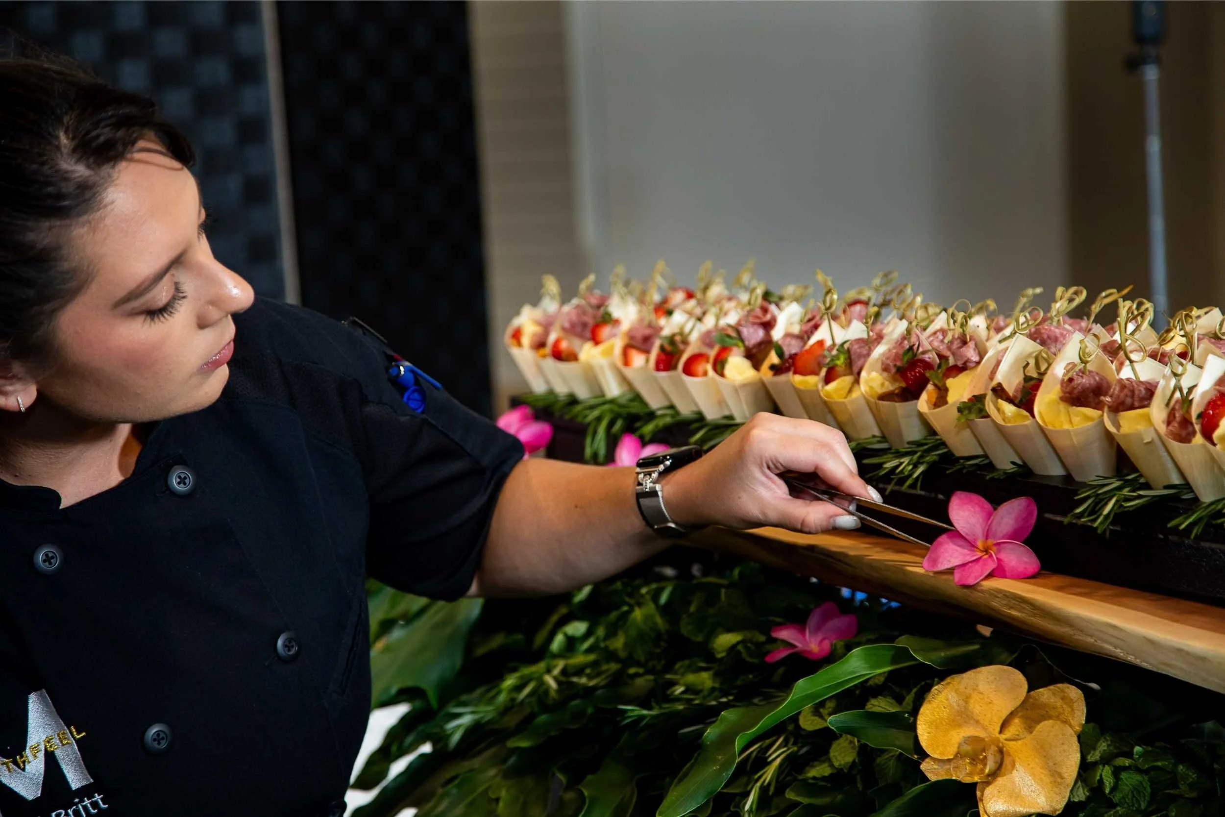 A woman in a black uniform arranging colorful appetizers on a table decorated with pink orchids and tropical leaves.