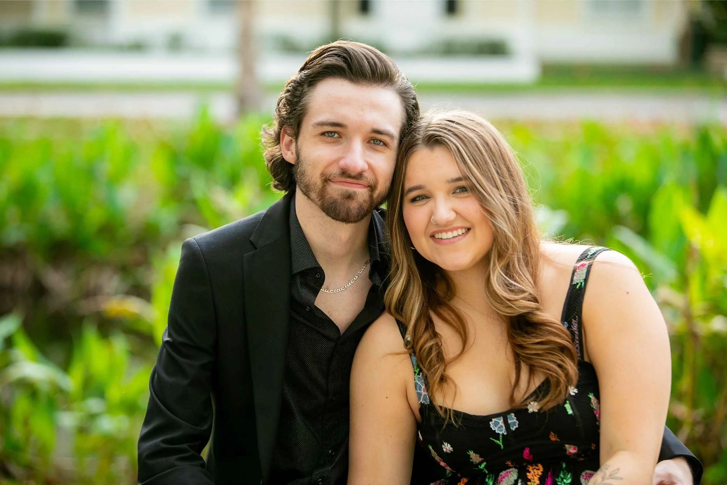 A smiling young man and woman sitting outdoors surrounded by greenery, close together and looking at the camera for an engagement photography session with Cannonfire Photography.