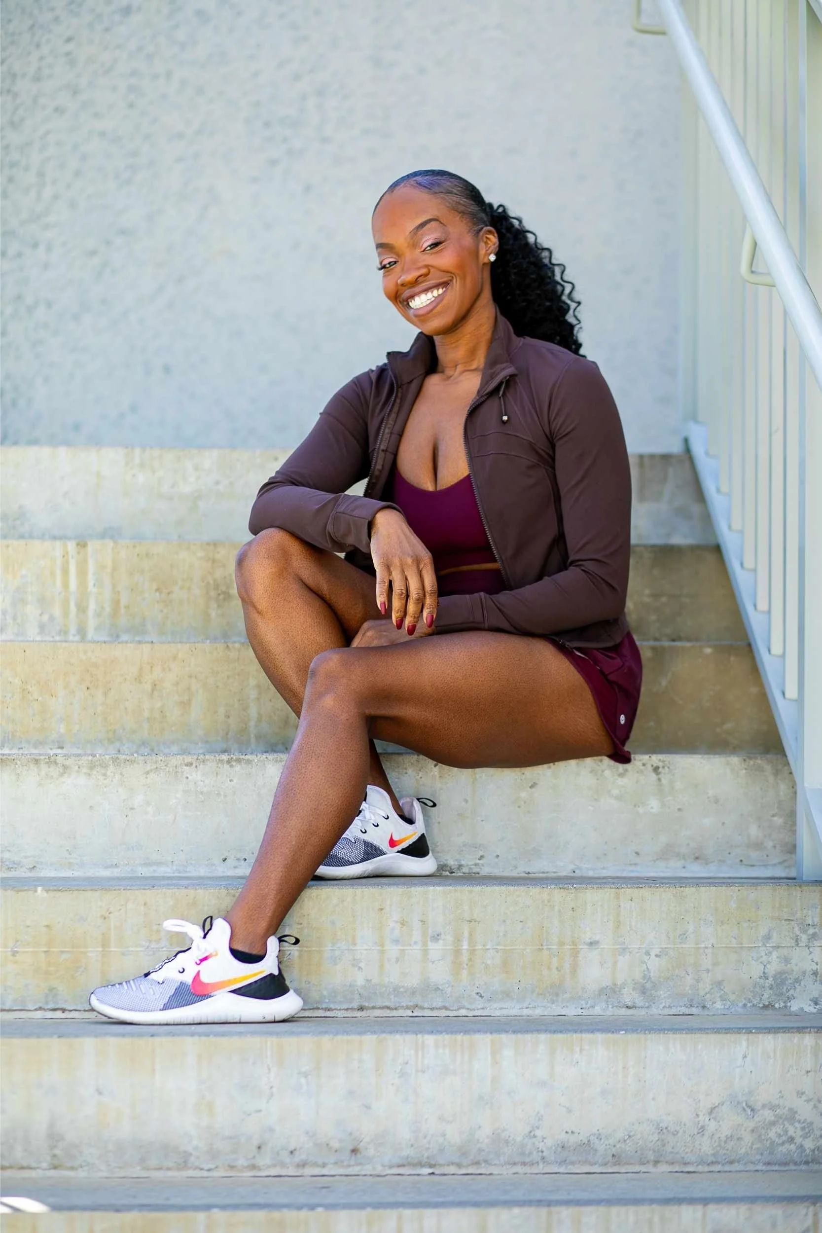A woman sitting on concrete stairs outside, smiling, wearing a brown jacket, maroon shorts, and Nike sneakers for a brand session with Cannonfire Photography.