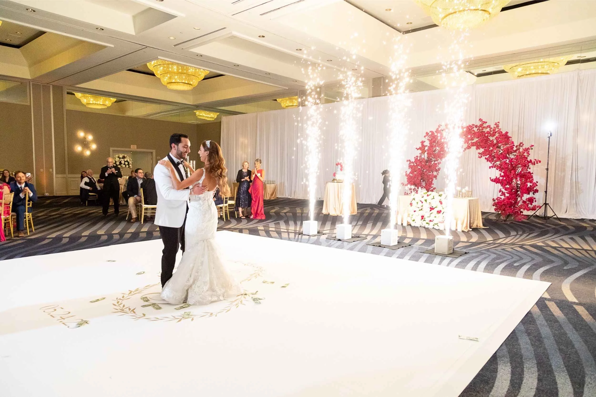 Bride and groom dancing at their wedding reception with sparklers and fireworks in the background, guests seated and standing around them, decorated with pink flowers and white drapes, for a wedding photographed by Cannonfire Photography.
