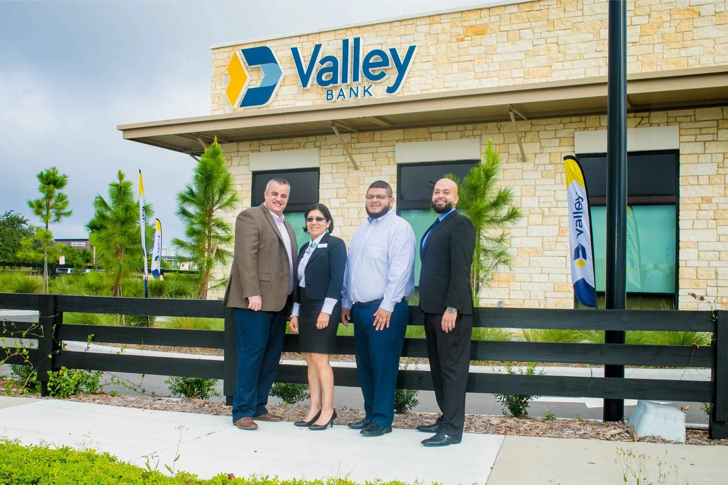 Group of four people standing in front of Valley Bank building with logo, trees, and flags for a team photo session by Cannonfire Photography.