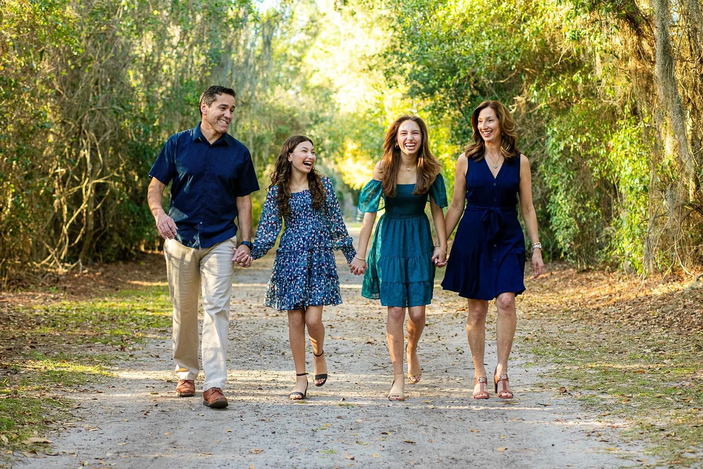 A family of four walking hand-in-hand on a dirt path through a wooded area, smiling and enjoying each other's company on a sunny day.
