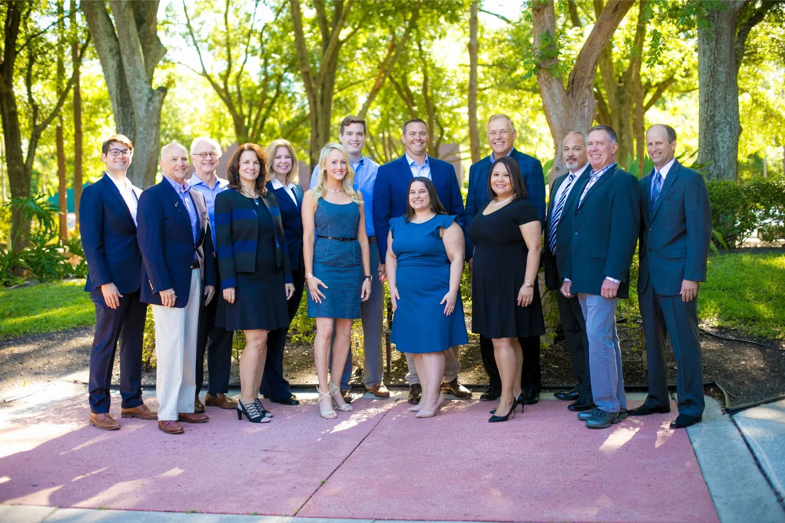 Group of professional men and women dressed in business attire, standing outdoors on a pink brick sidewalk in front of trees with green leaves for a team photo by Cannonfire Photography.