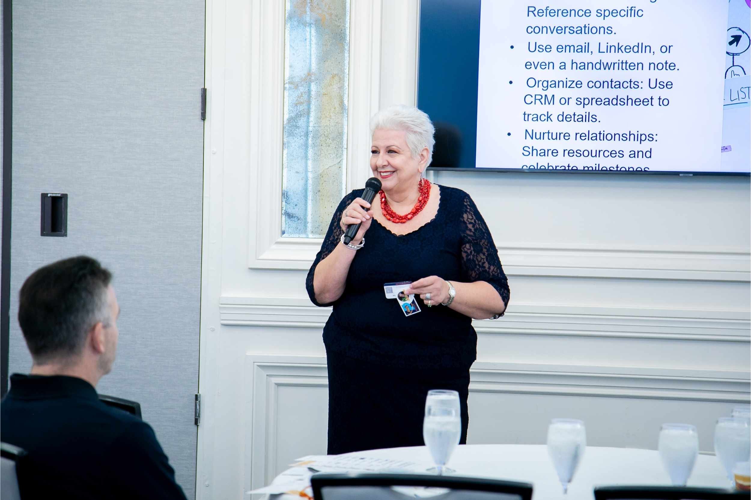 An older woman with short white hair and a red necklace is giving a presentation using a microphone. She is standing in front of a large screen displaying text, with a woman and a man seated at a table with glasses of water and a laptop.