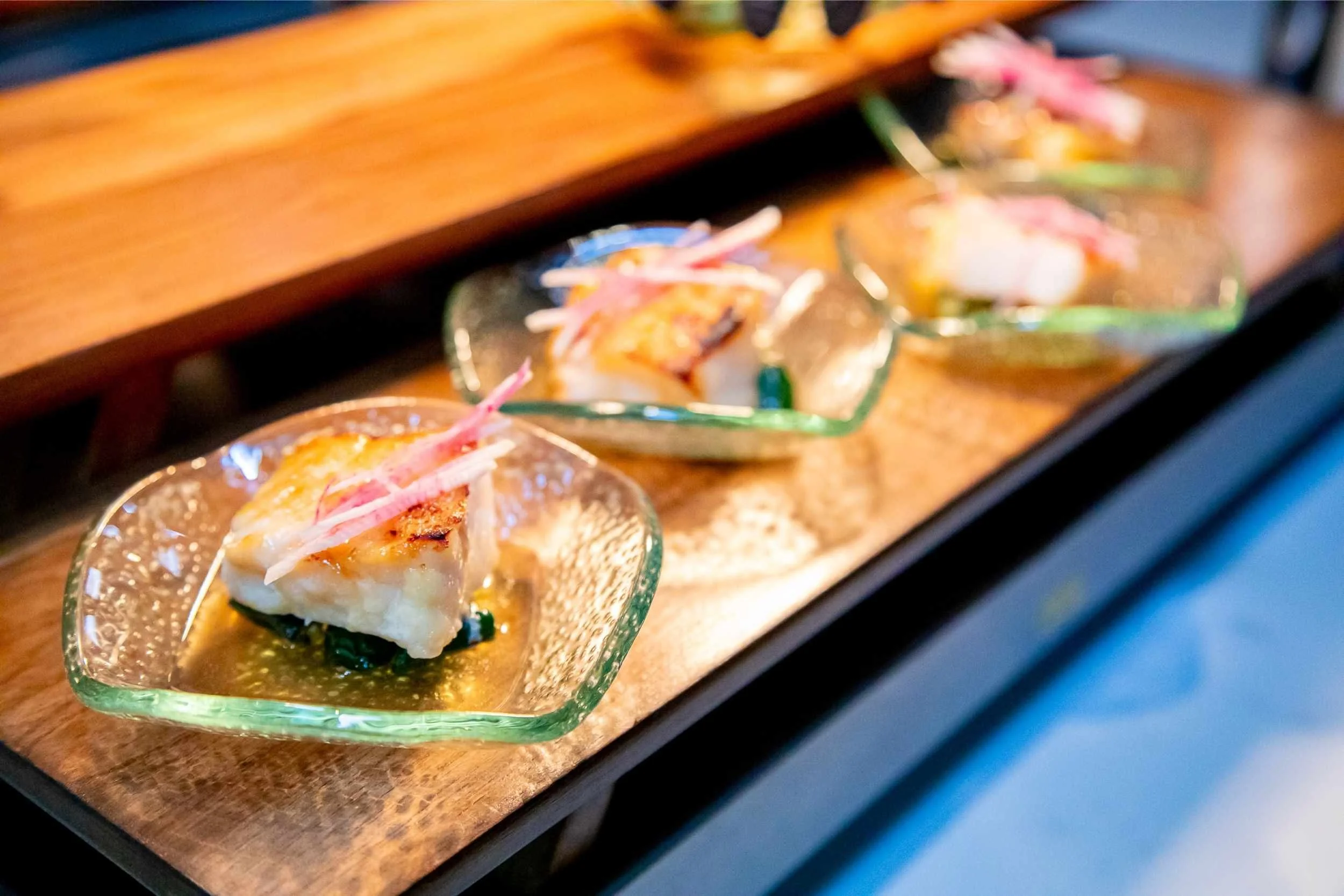 Three plated pieces of gourmet fish with garnishes and sauce on a wooden serving board.