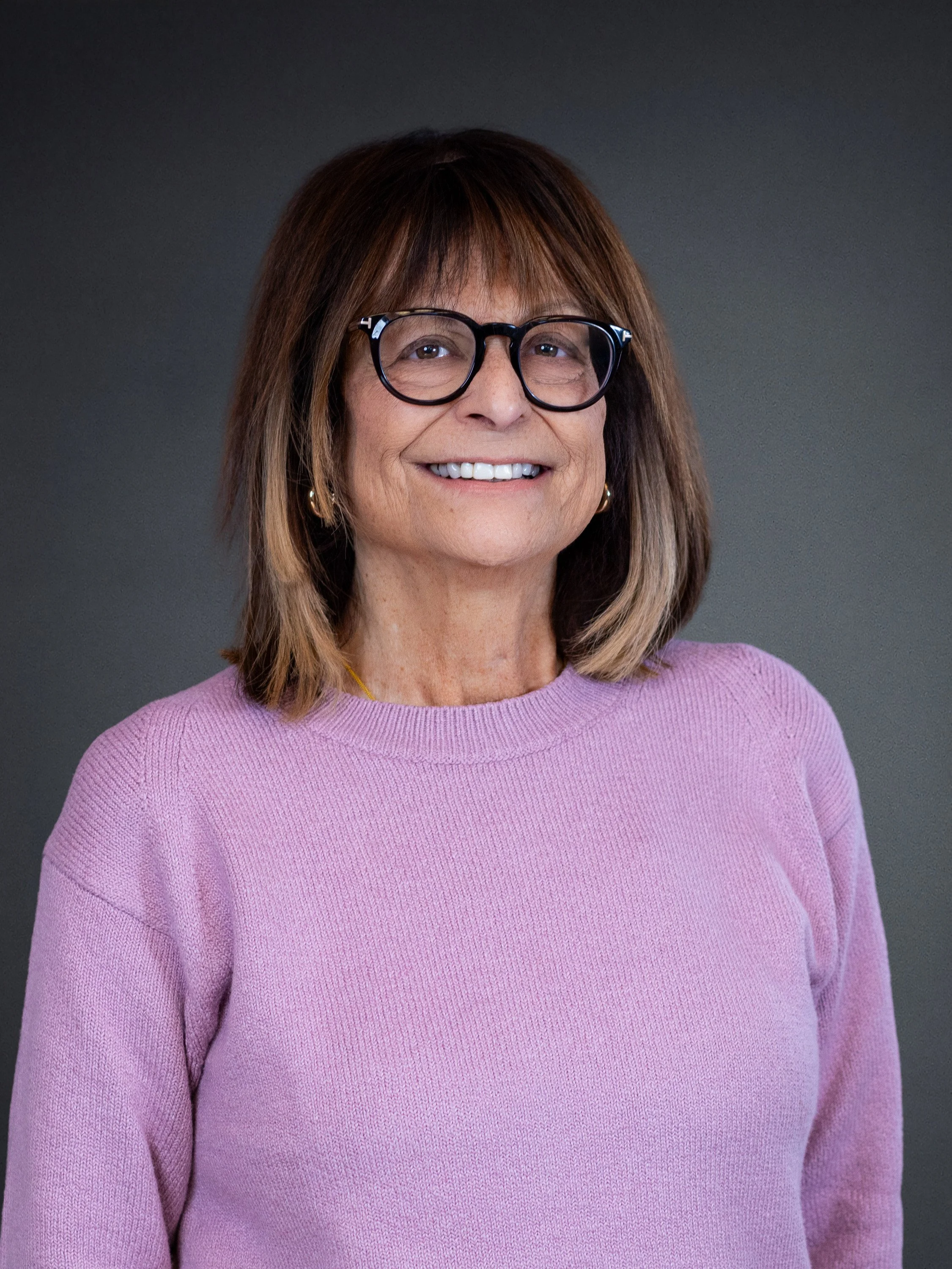 A smiling woman with shoulder-length brown hair, wearing glasses and a pink sweater. She is standing against a dark gray background for a corporate headshot with Cannonfire Photography in Orlando.