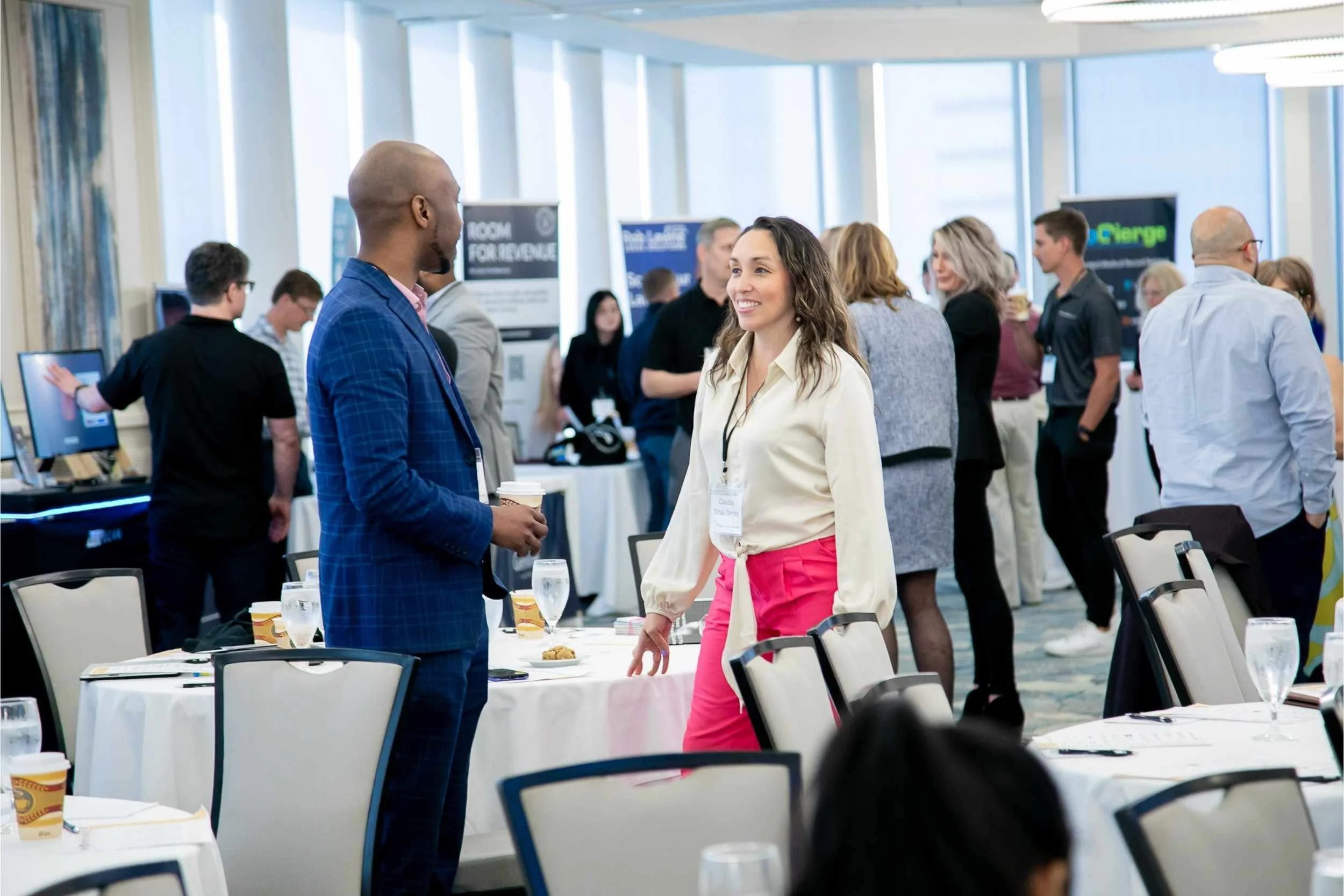 A conference room with several people engaging in conversations, some standing and some sitting at tables set with glasses and coffee cups.