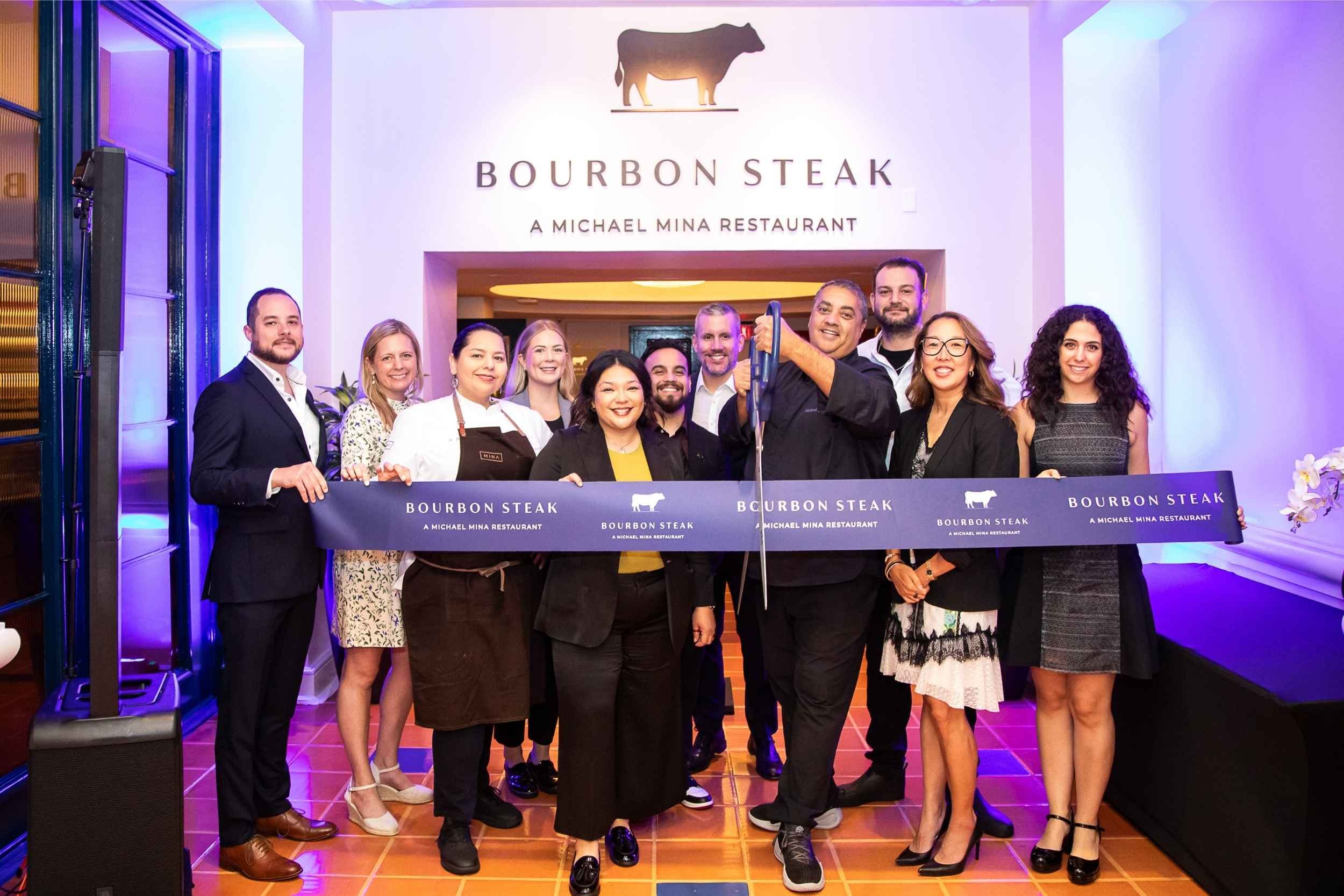 Group of people at a restaurant ribbon-cutting event in front of a sign that says "Bourbon Steak, A Michael Mina Restaurant," with all smiling and holding a ribbon that also displays the restaurant's name.