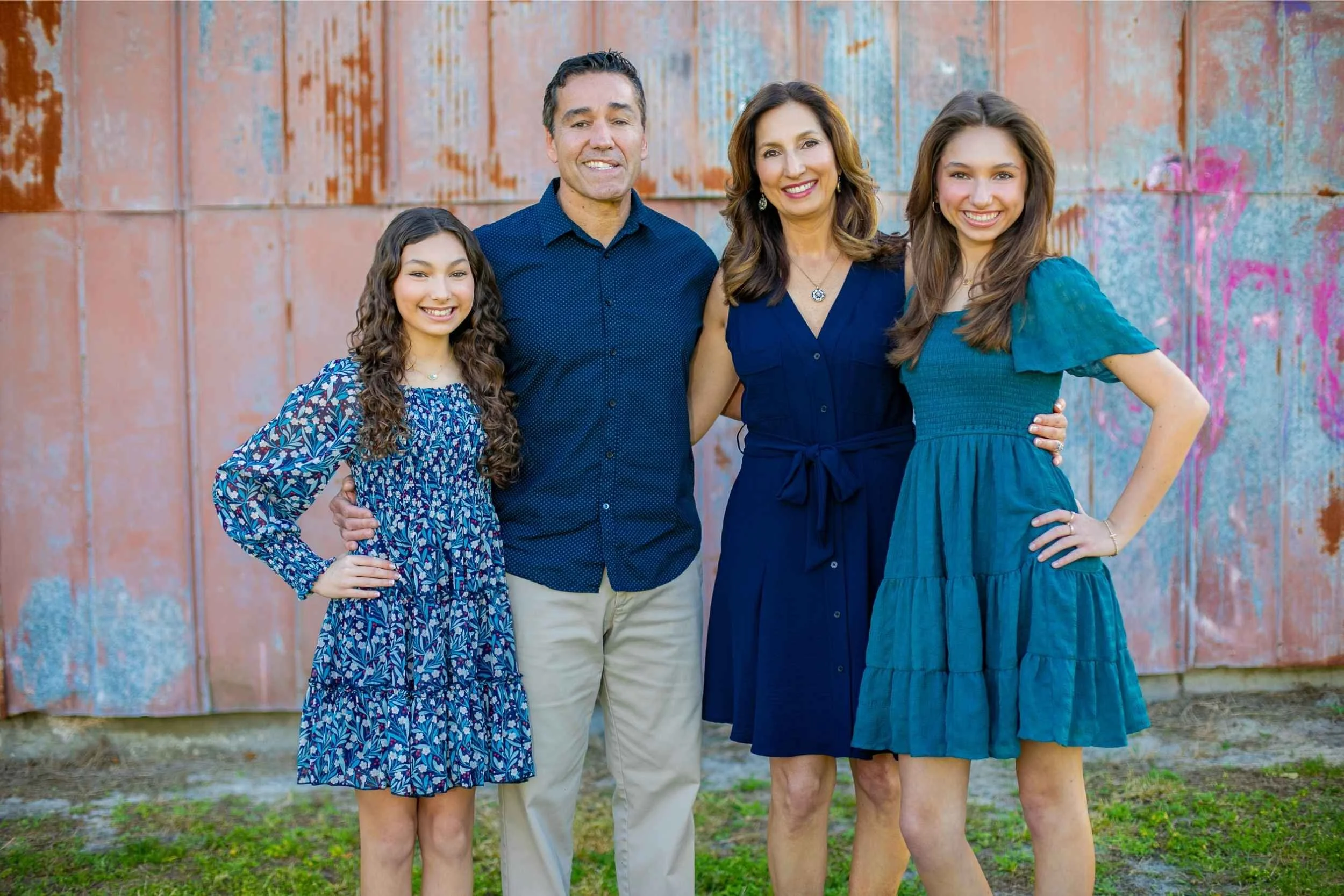 A family of four standing outdoors in front of a rusted metal wall, smiling at the camera. One adult woman and two girls with long brown hair, and a man with dark hair.