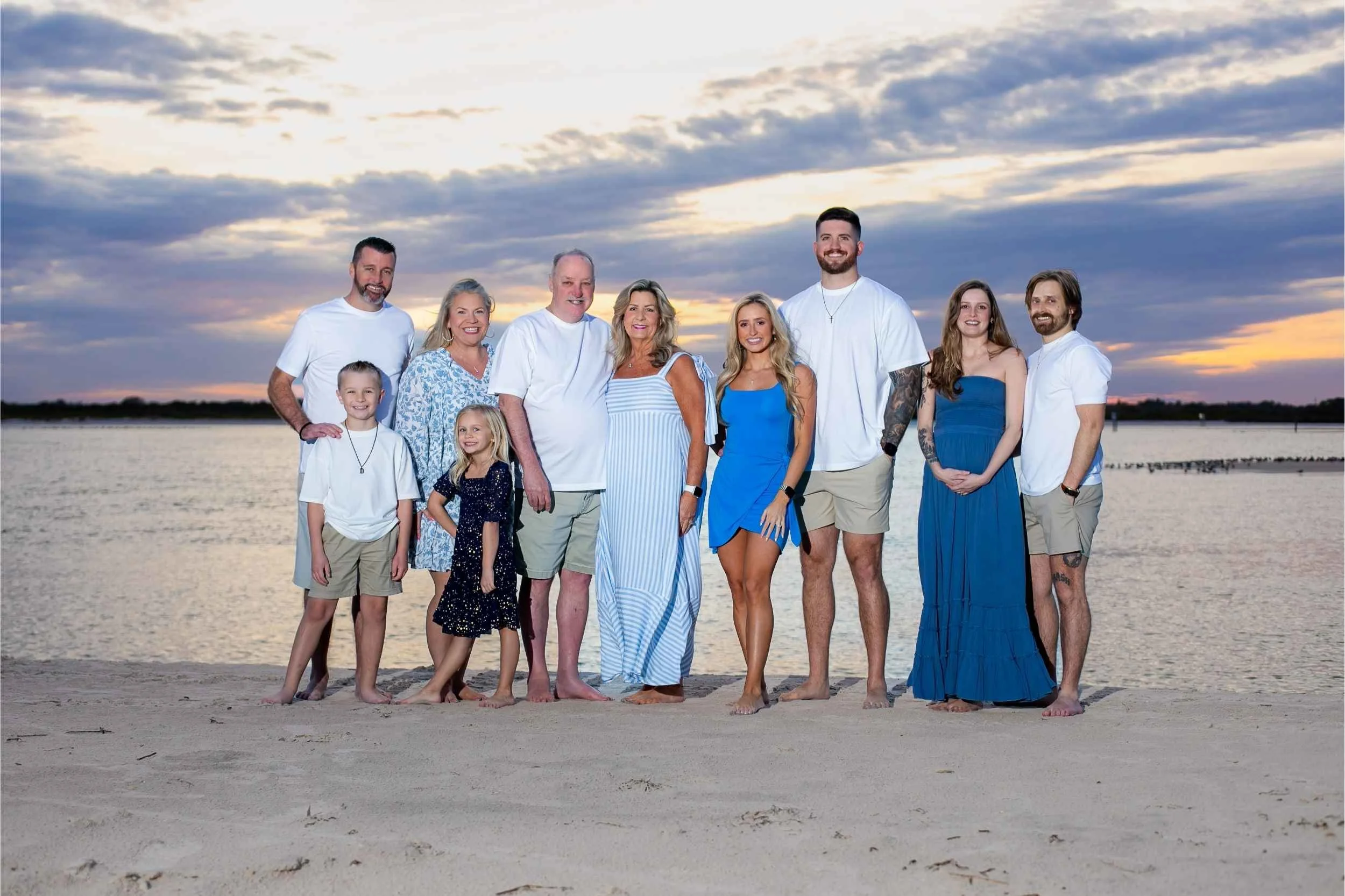 A multi-generational family group standing on a sandy beach near a body of water during sunset, smiling at the camera. The group includes children, adults, and seniors dressed in casual blue and white summer clothing.